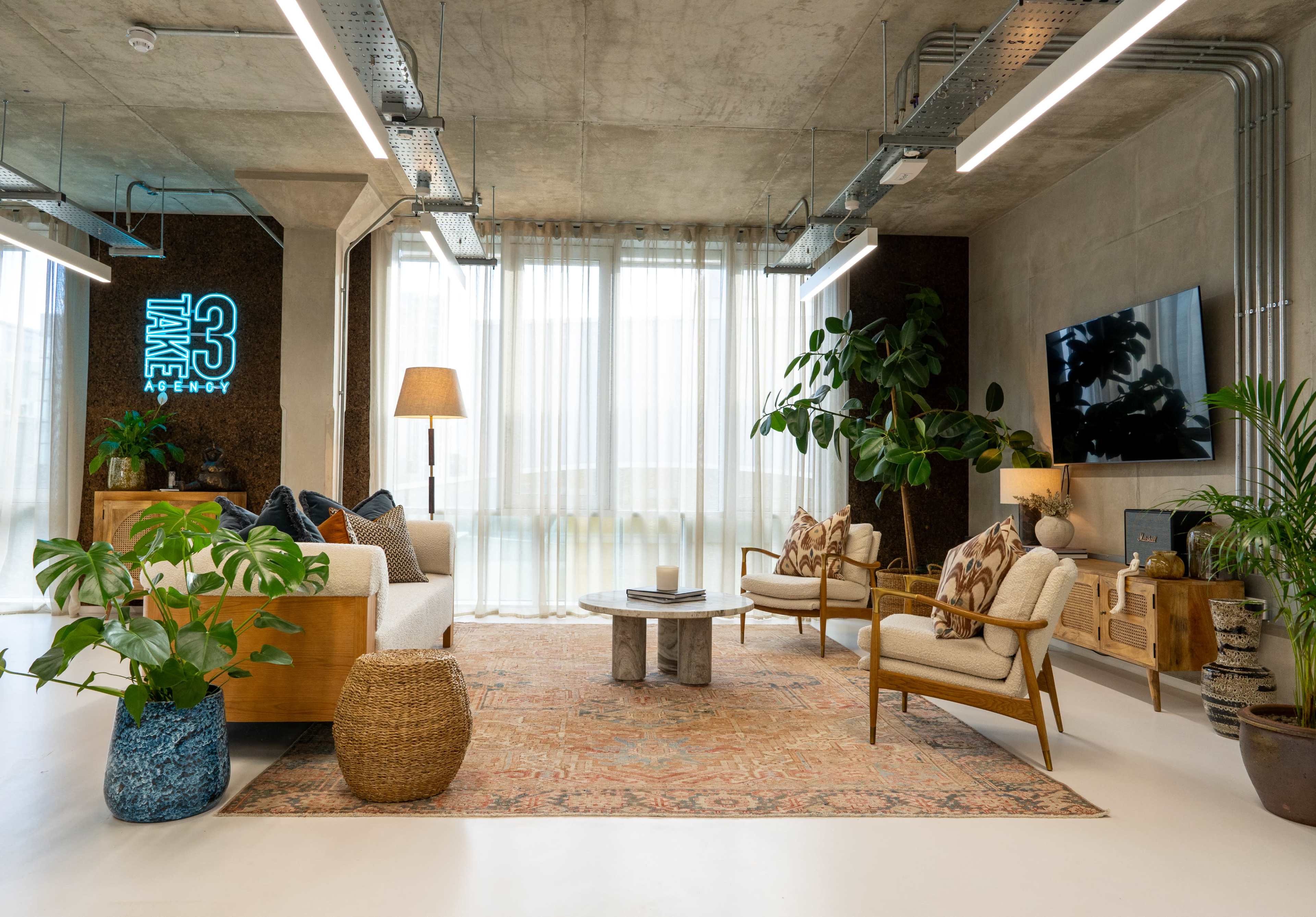 The image shows a modern living area with a light-colored sofa, armchairs, a wooden table, a large rug, and plants, illuminated by natural light filtering through sheer curtains.