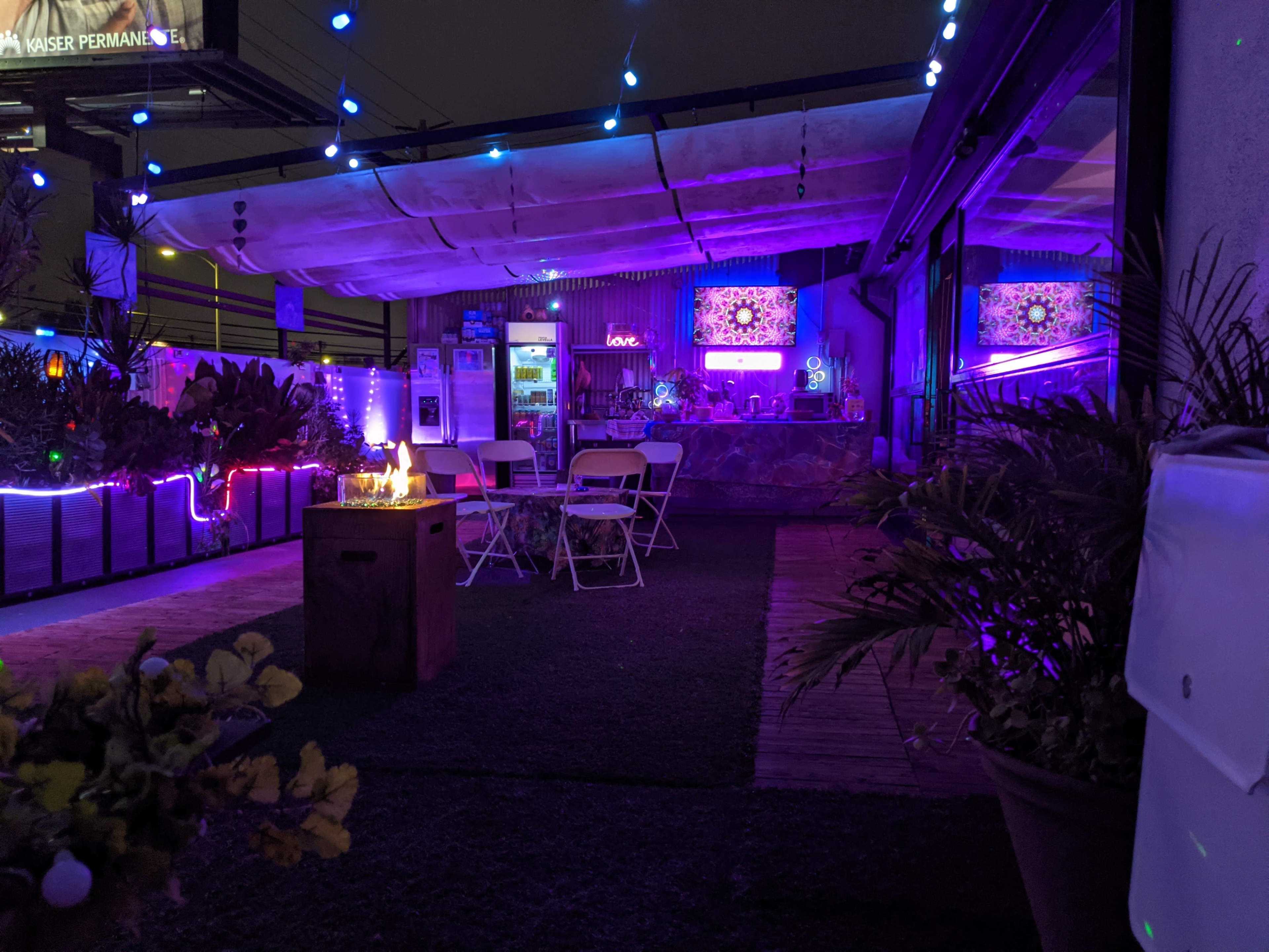 The image shows a rooftop space adorned with colorful lights, plants, and chairs, creating a vibrant evening atmosphere.