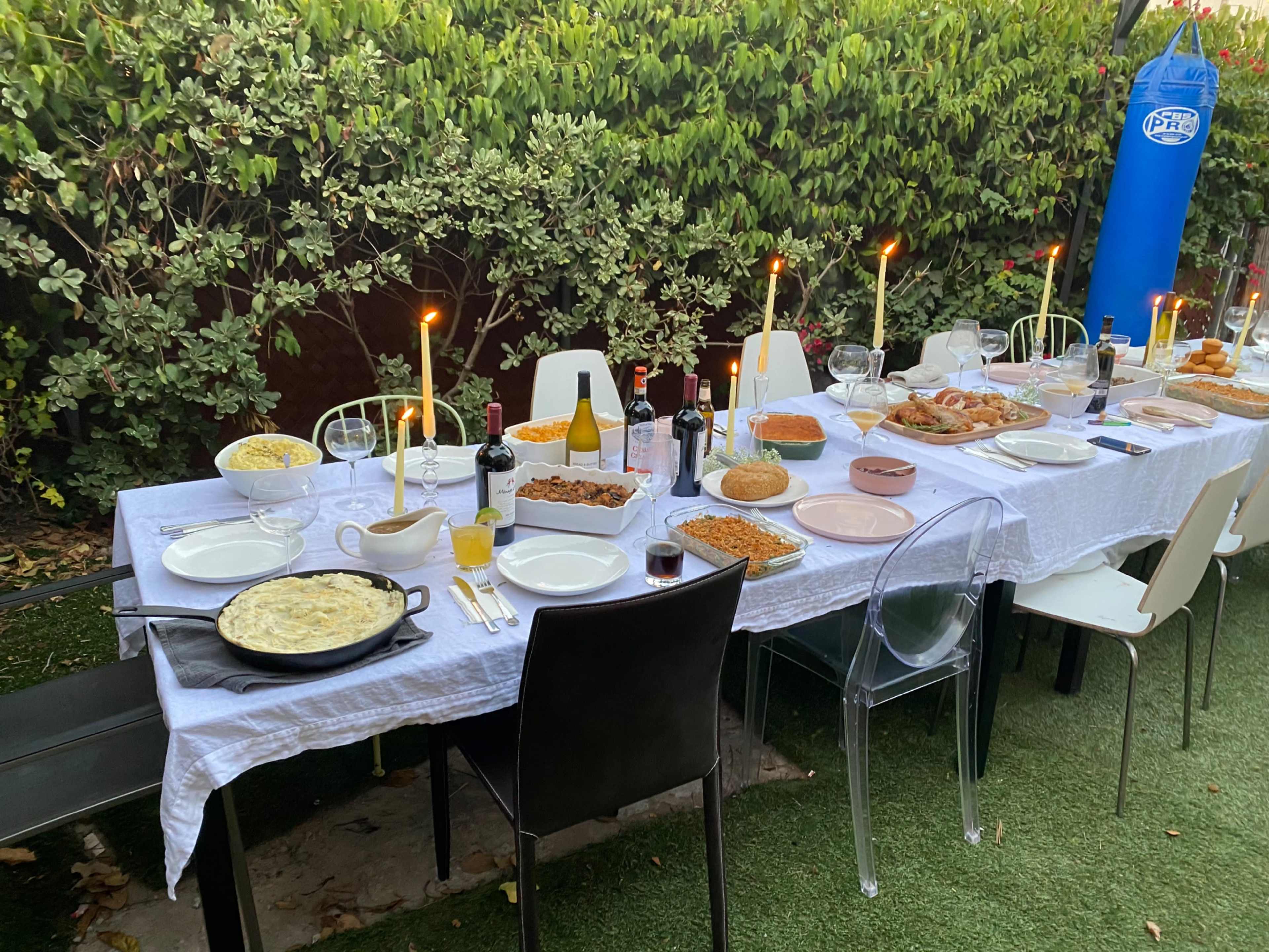 A long dining table is set up outdoors, covered with a white tablecloth and surrounded by chairs, featuring various dishes, bottles, glasses, and lit candles.