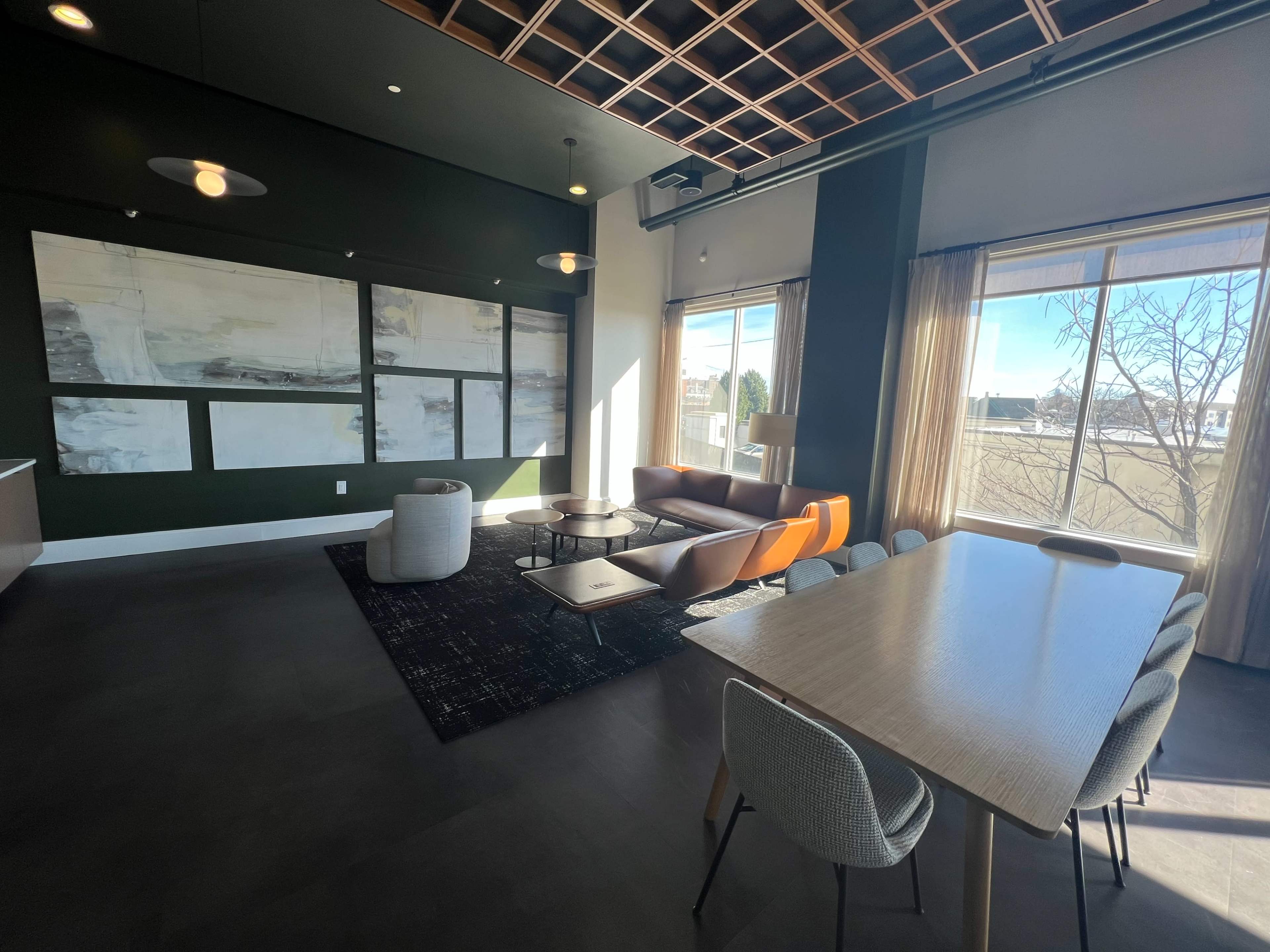 A modern lounge area featuring a seating arrangement with a dark rug, a large table, and large windows allowing natural light to enter.
