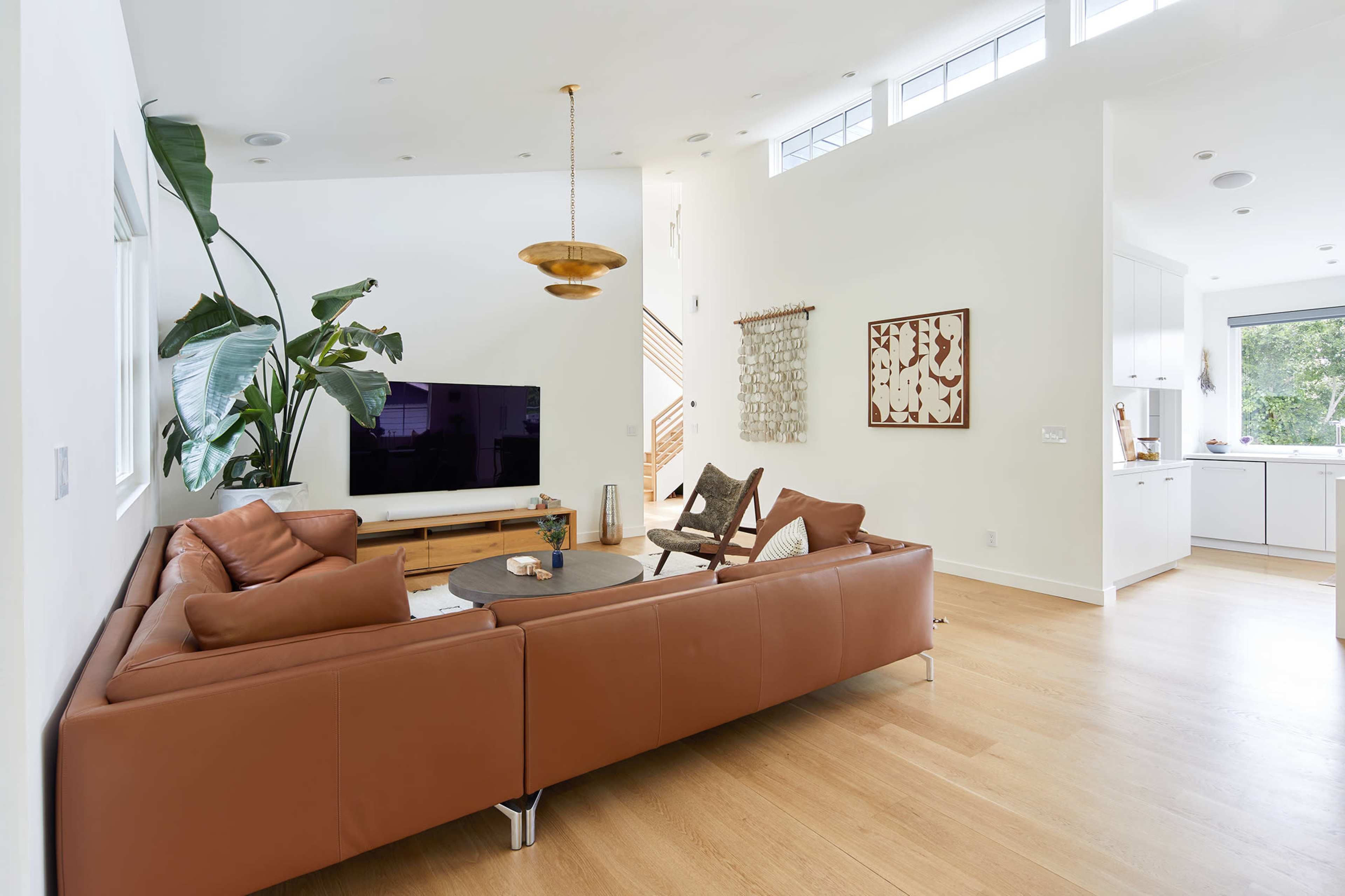 A modern living room features a brown leather sofa, a round coffee table, a large TV, and a well-lit kitchen area in the background.