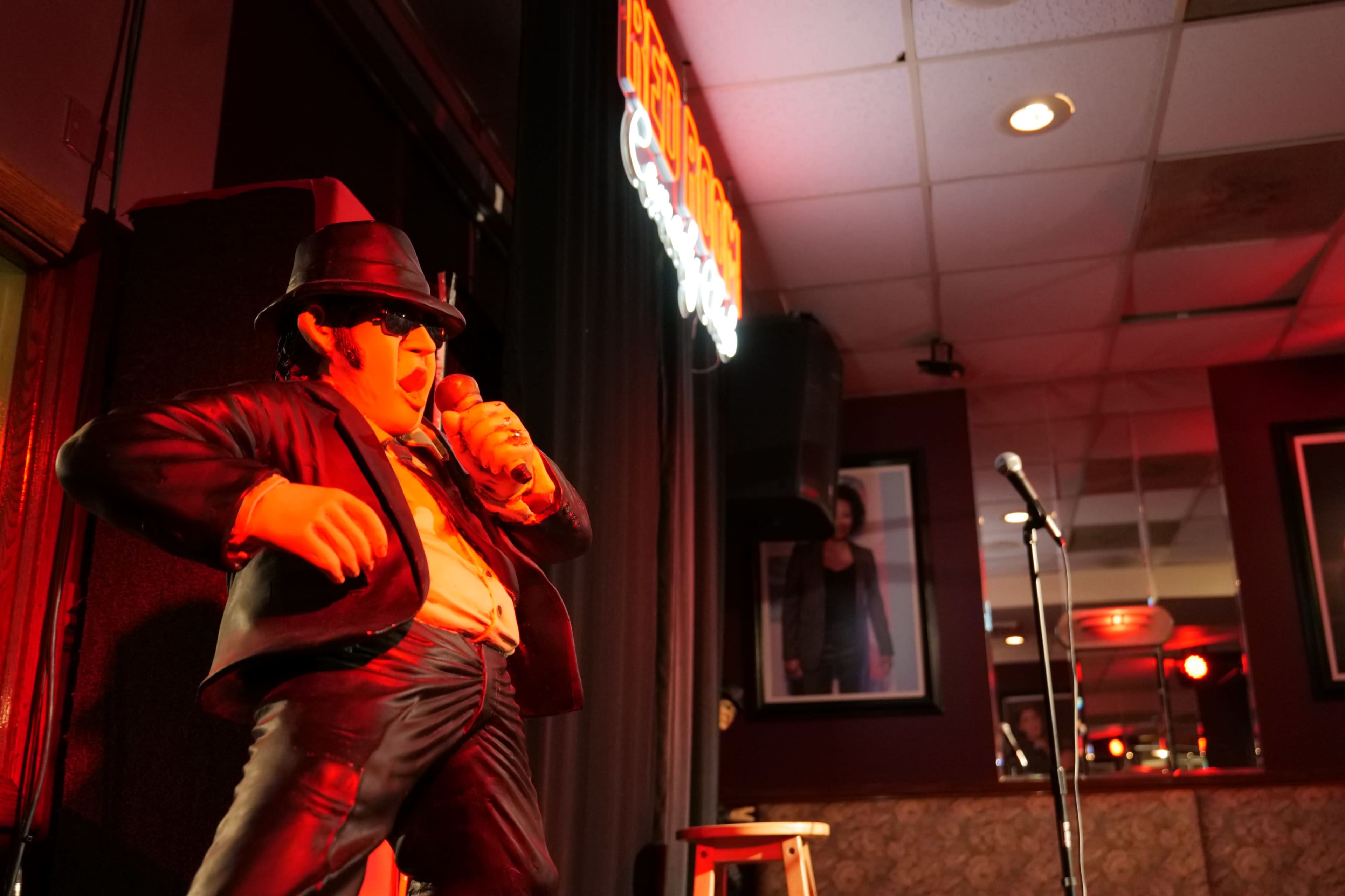 A life-sized statue of a man in a black suit and hat stands on a stage, holding a microphone with a spotlight directed at him, while a dimly lit bar background features a microphone stand and a neon sign.