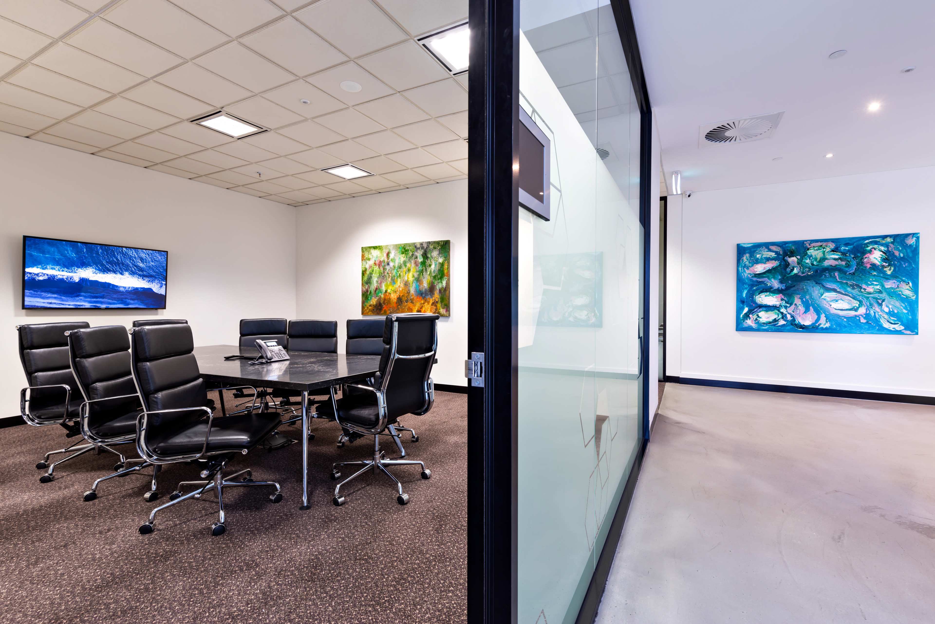 A modern conference room features a large table surrounded by black leather chairs, with two colorful paintings on the adjacent walls.