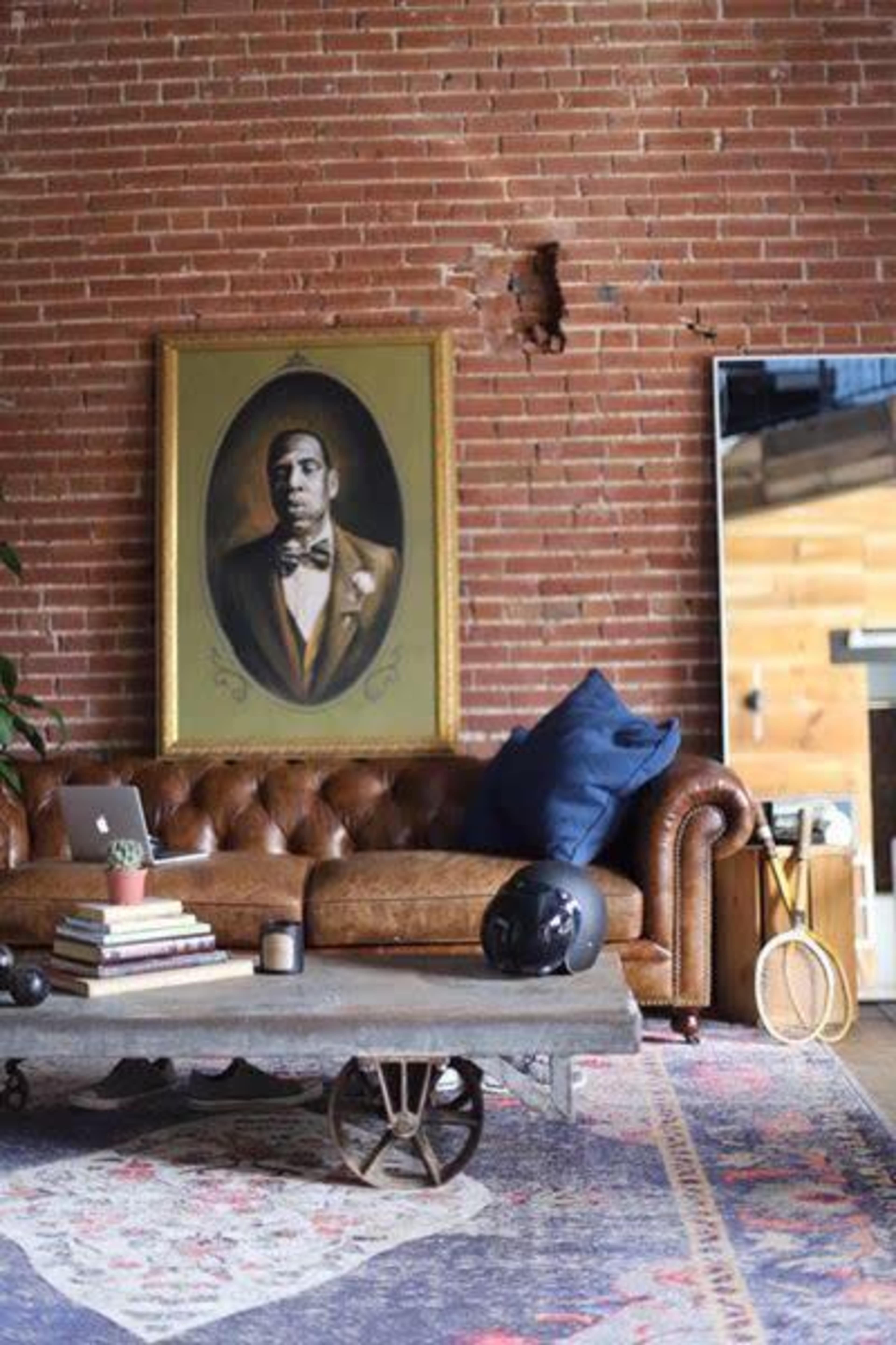 The image features a leather sofa against a brick wall, with a portrait of a man in formal attire hanging above it, and a rustic coffee table adorned with books and a decorative item.