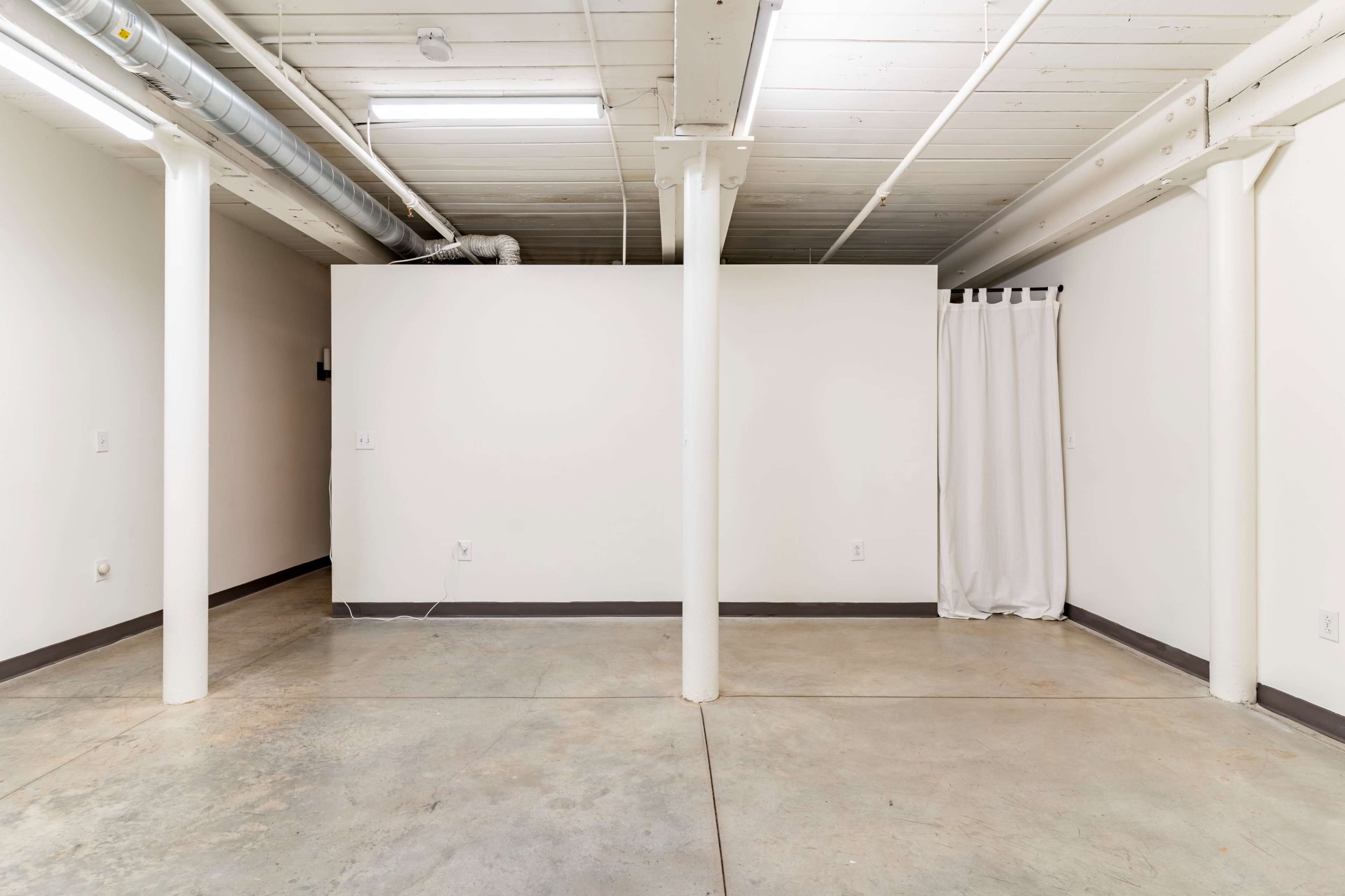 An empty basement room with concrete flooring, white walls, and a curtain in the corner.