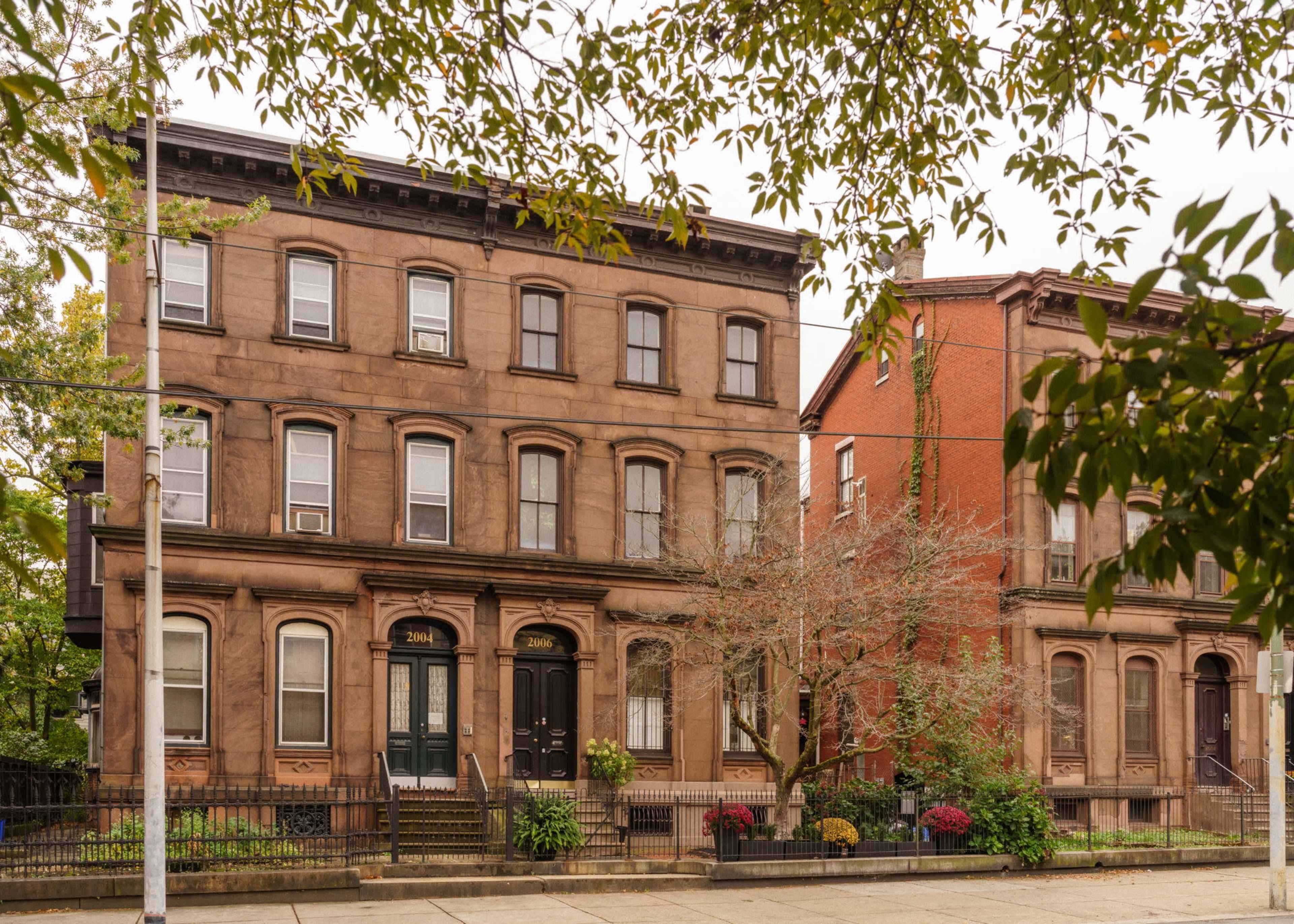 The image shows two historic brick apartment buildings, one brownstone and the other red, set on a tree-lined street with planters and a small tree in front.