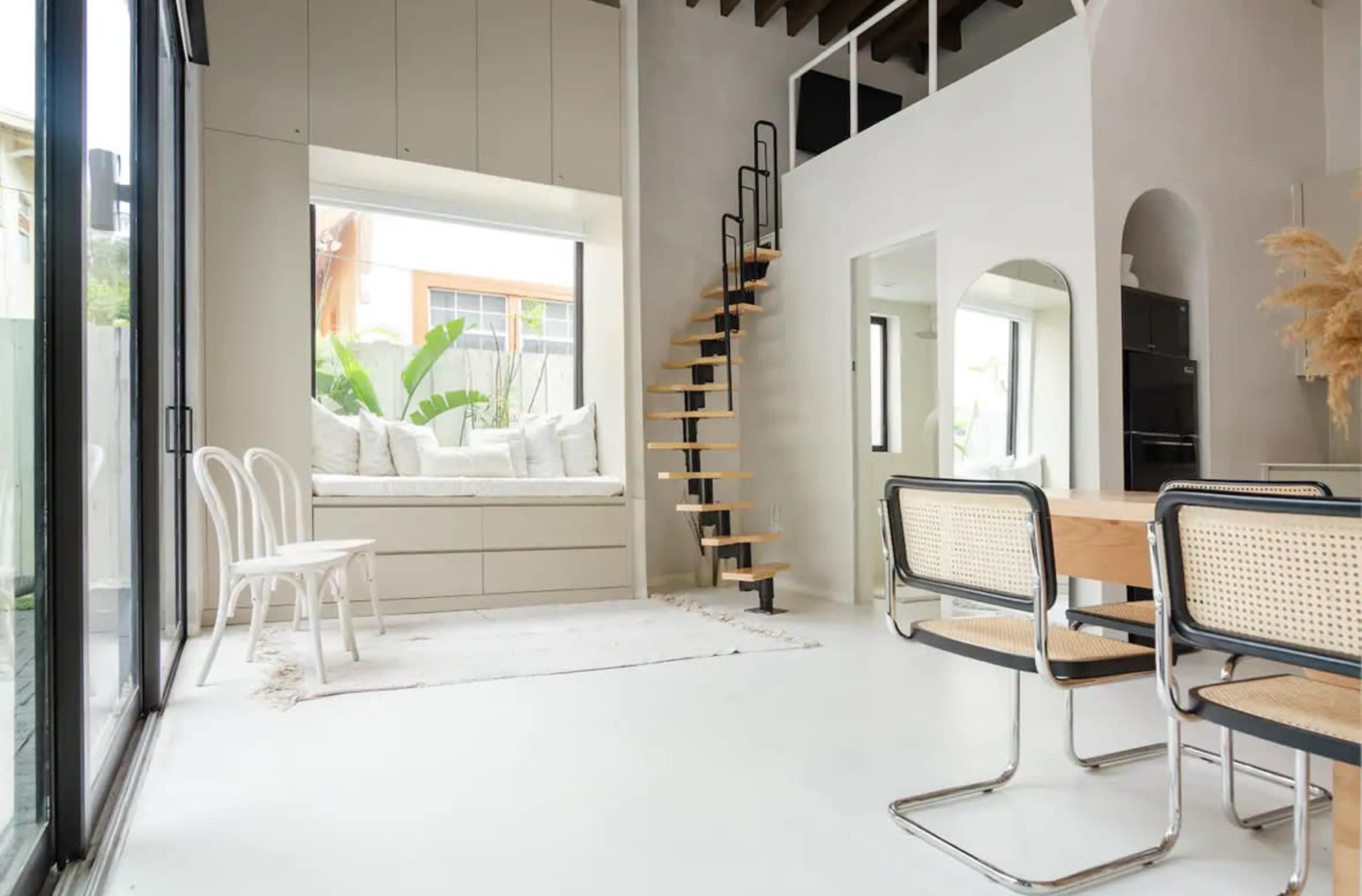 The image shows a modern living space featuring a spiral staircase, a dining area with chairs, and a large window with a built-in seating nook.