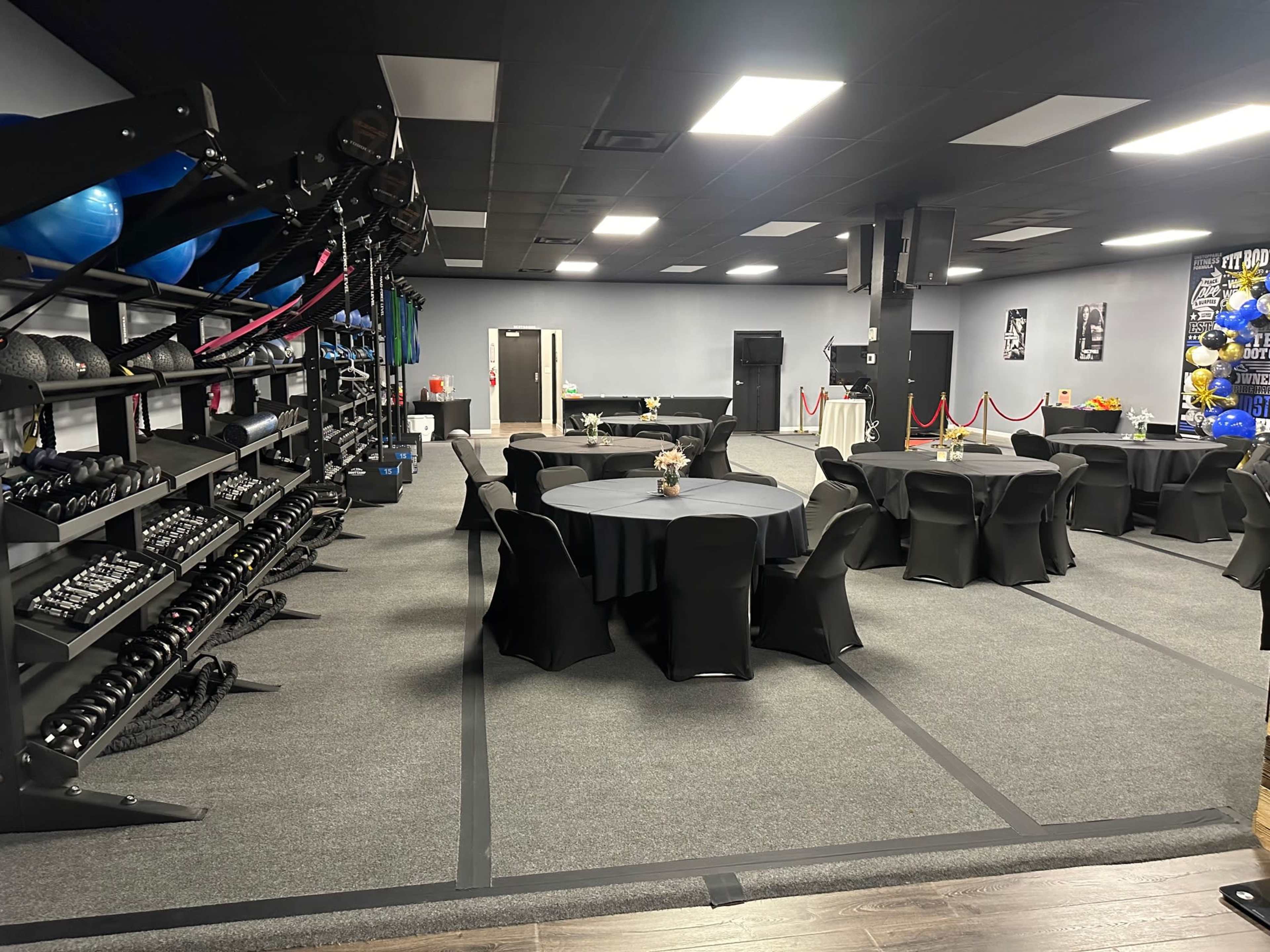 The image shows an indoor event space featuring several round tables with black tablecloths and a fitness equipment area equipped with weights and exercise balls.