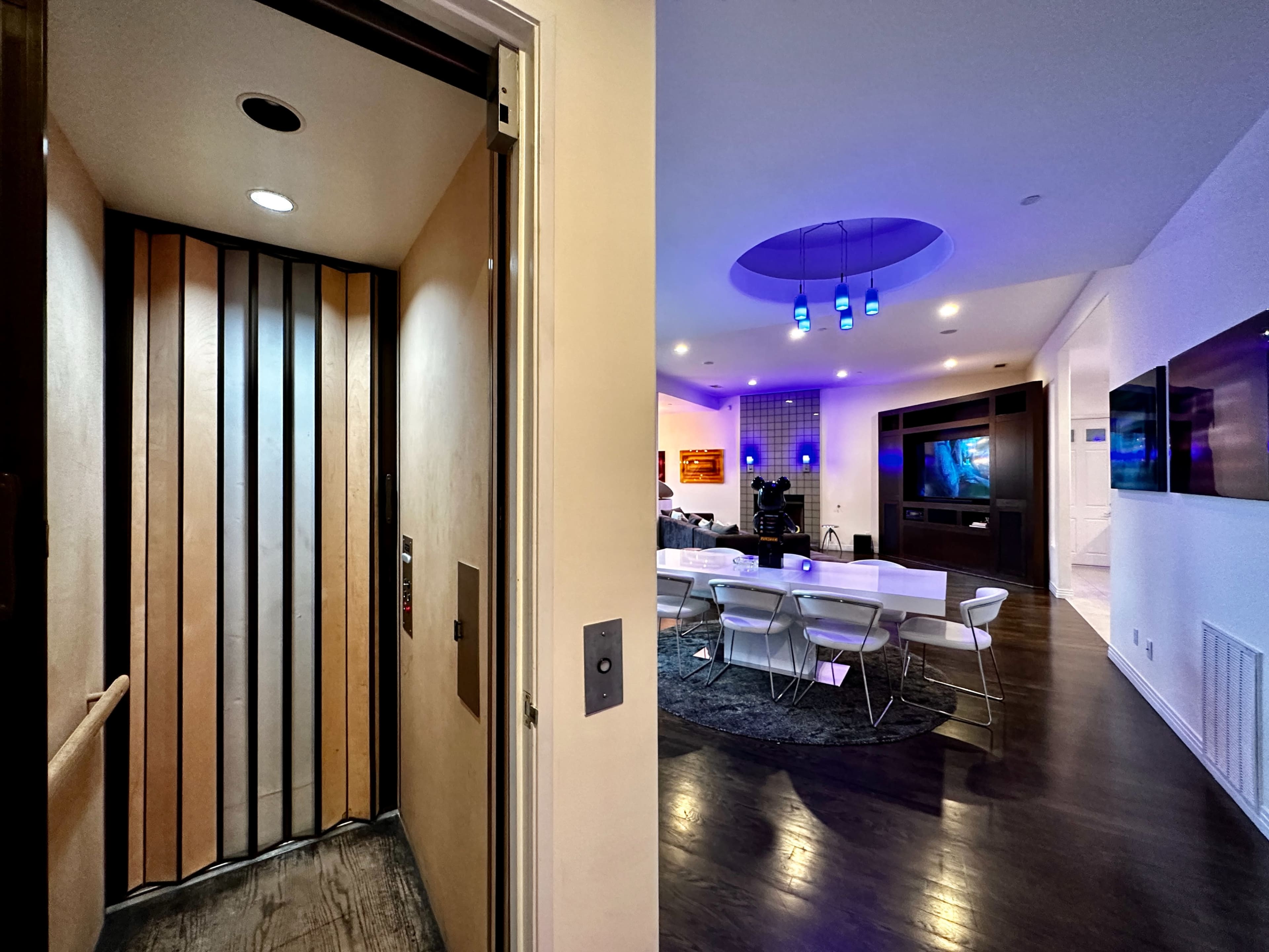 The image shows an elevator on the left and a modern living space with a dining area and entertainment setup on the right.