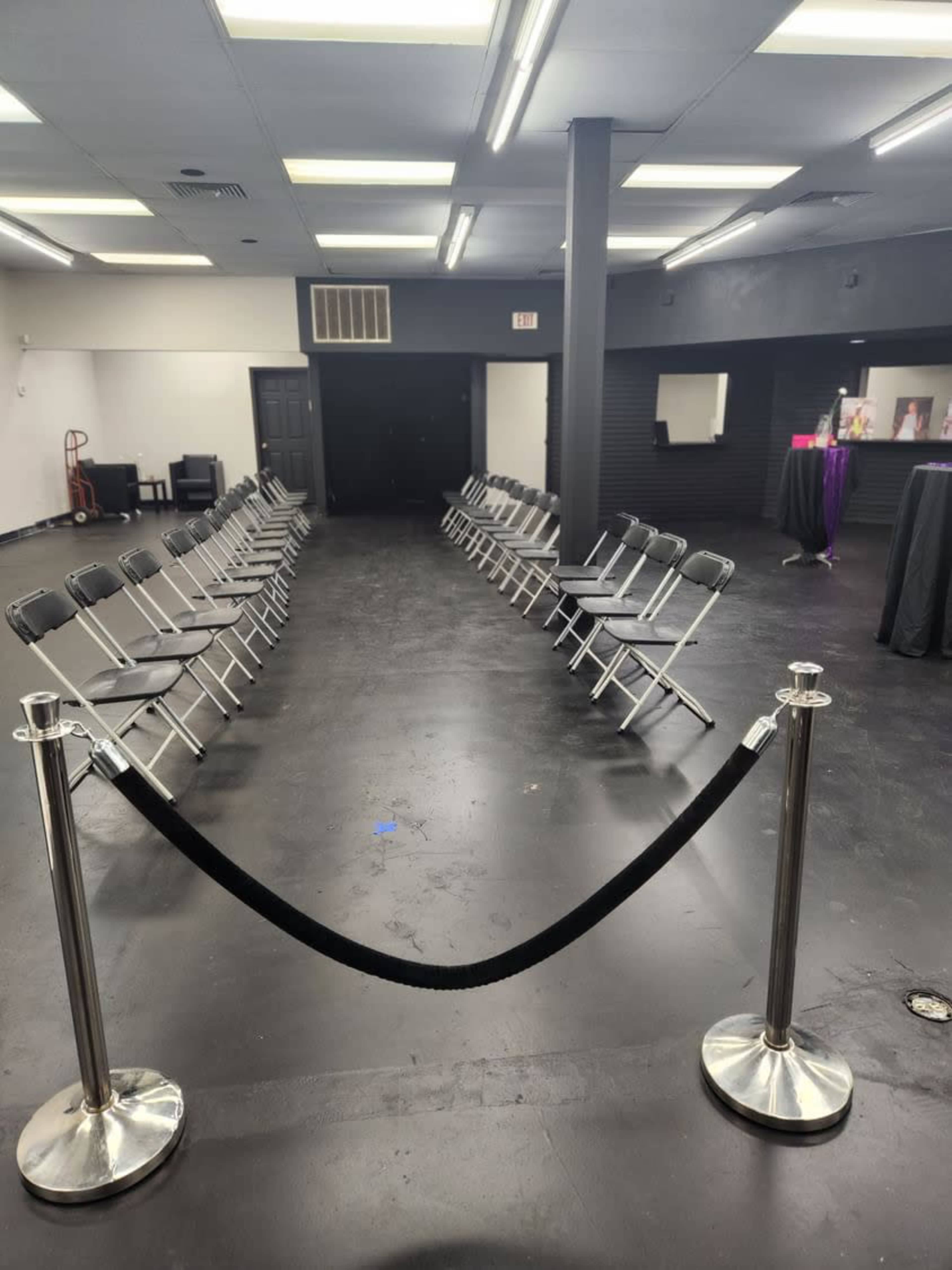 A row of metal chairs is arranged in two lines facing each other in a spacious, empty room with black flooring and bright overhead lights.