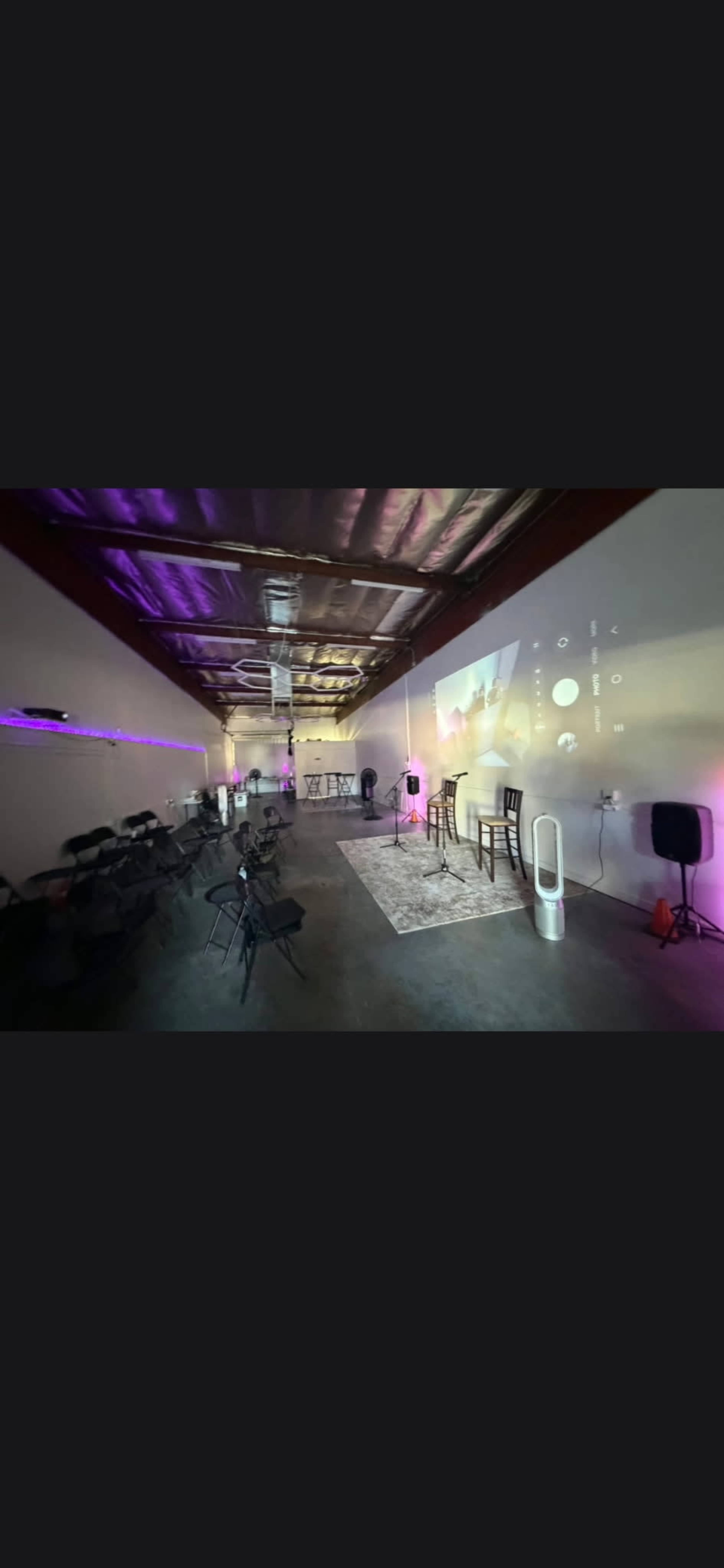 The image shows an empty indoor event space with chairs arranged for a presentation, ambient lighting, a projector displaying information on a wall, and audio equipment in the corners.