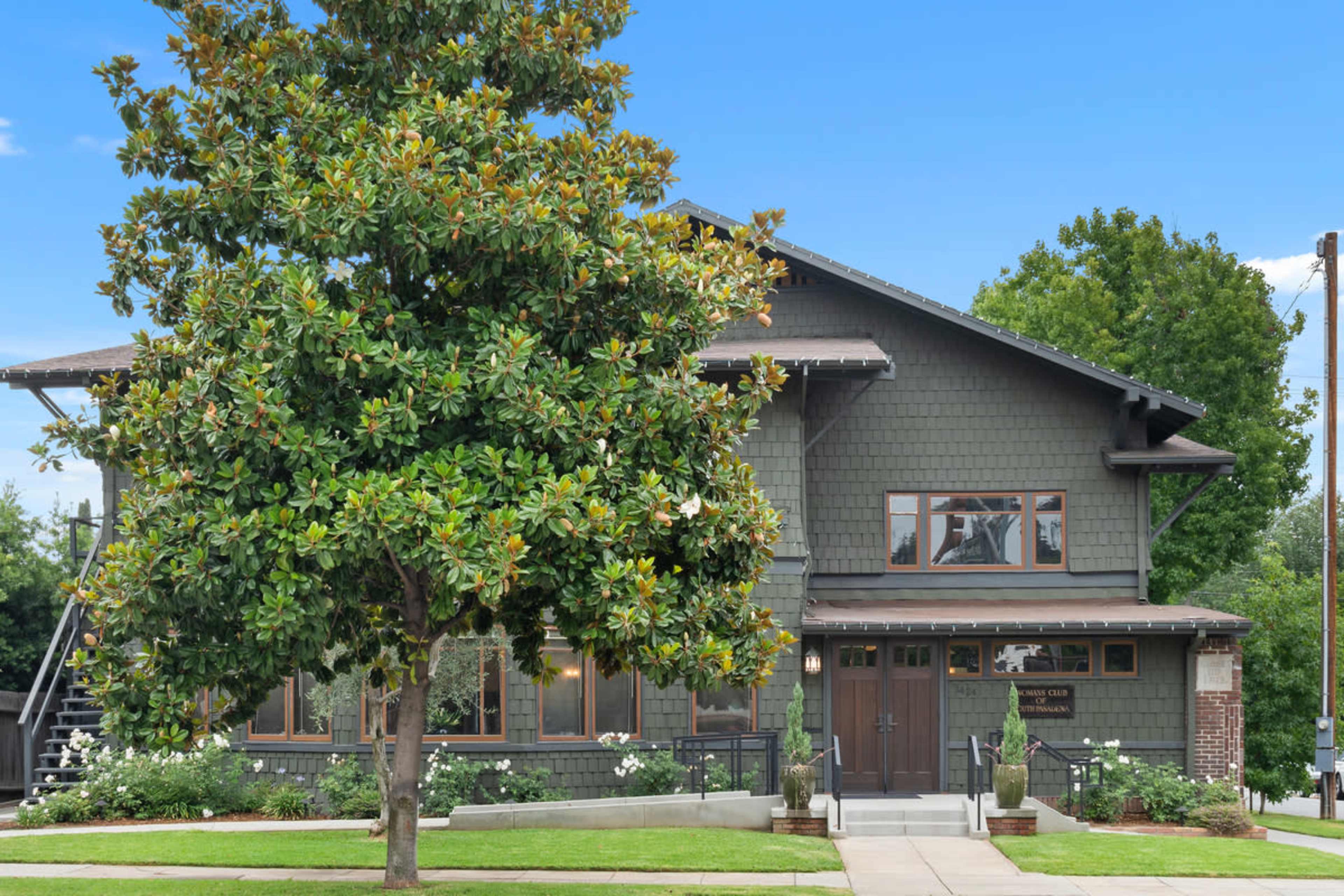 A two-story house with a dark exterior, large windows, and a front porch is surrounded by green grass and a magnolia tree.