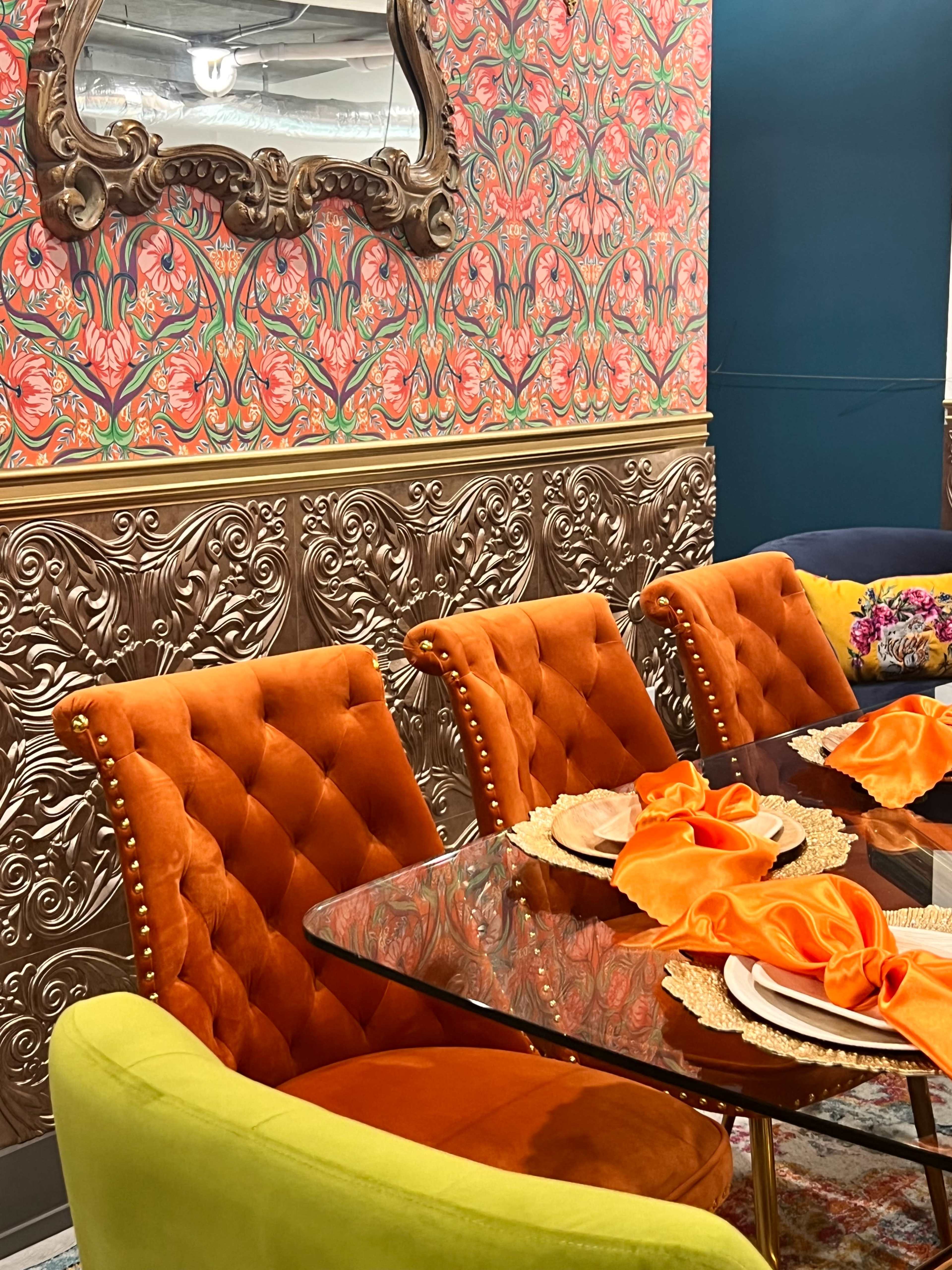 A dining area featuring a glass table with decorative plates and orange napkins, accompanied by orange upholstered chairs and a gold-accented wall.