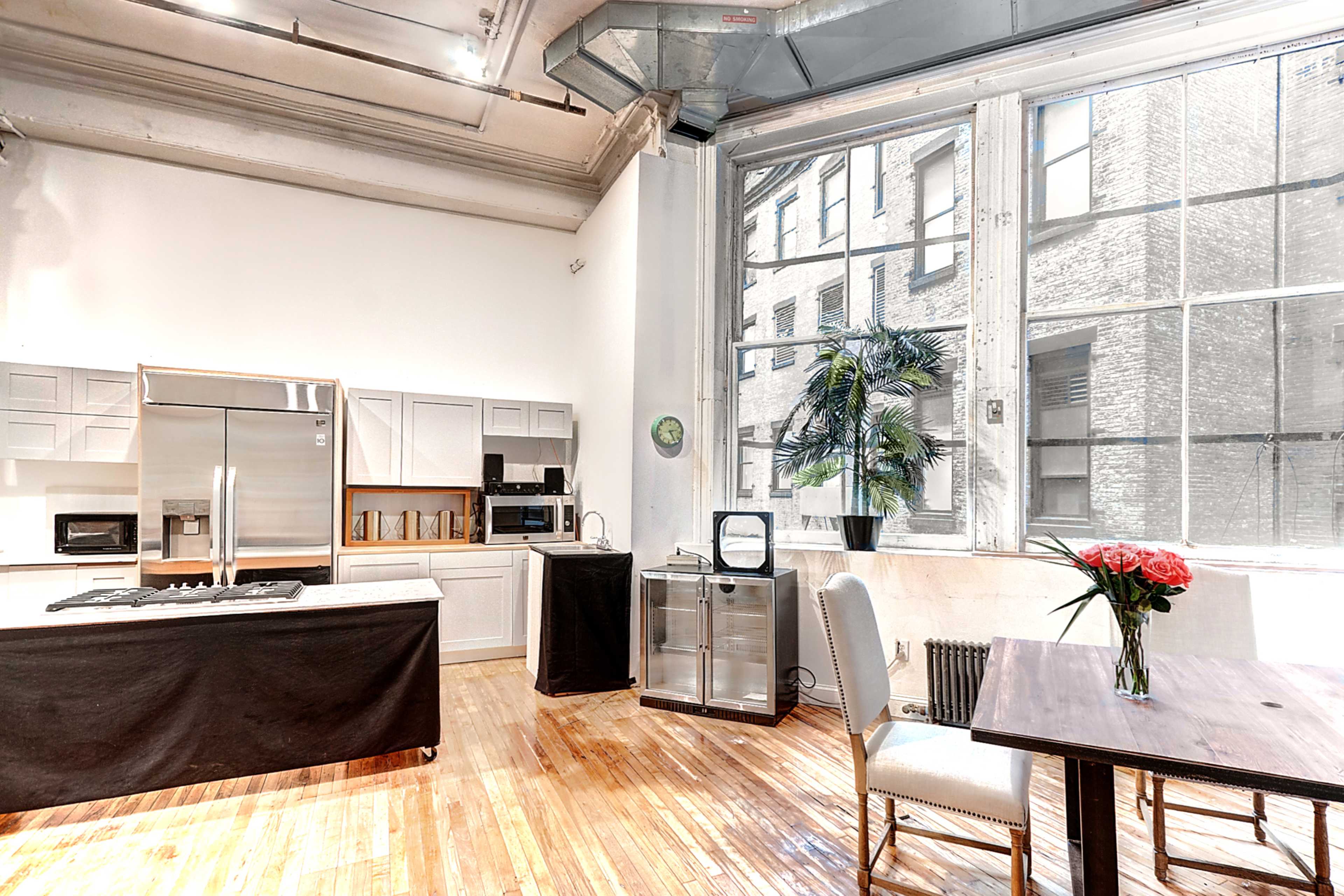 The image shows a spacious, airy kitchen-living area with large windows, hardwood floors, and modern appliances.