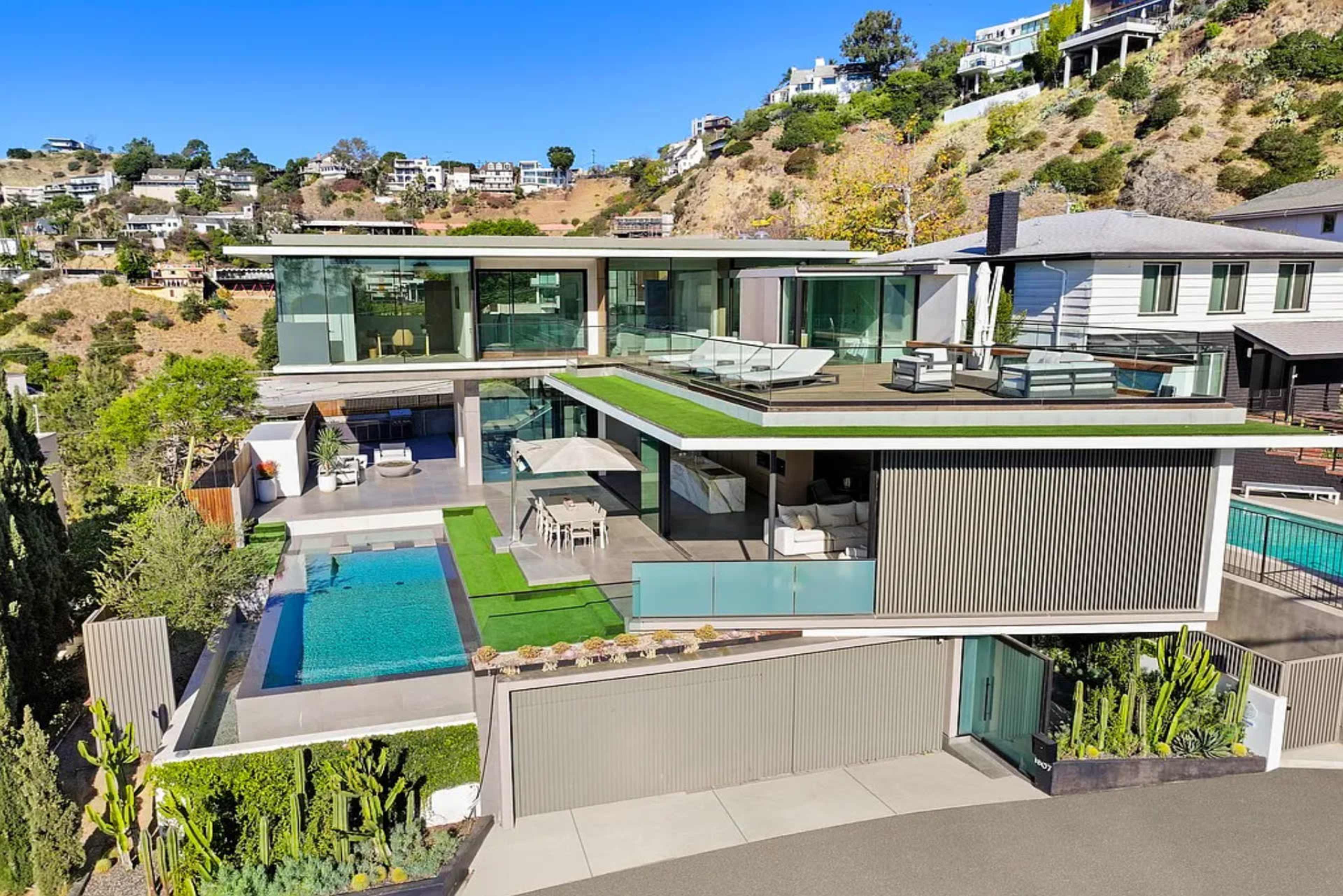 The image shows a modern, multi-level house with a flat roof, large glass windows, and an outdoor pool surrounded by green landscaping.