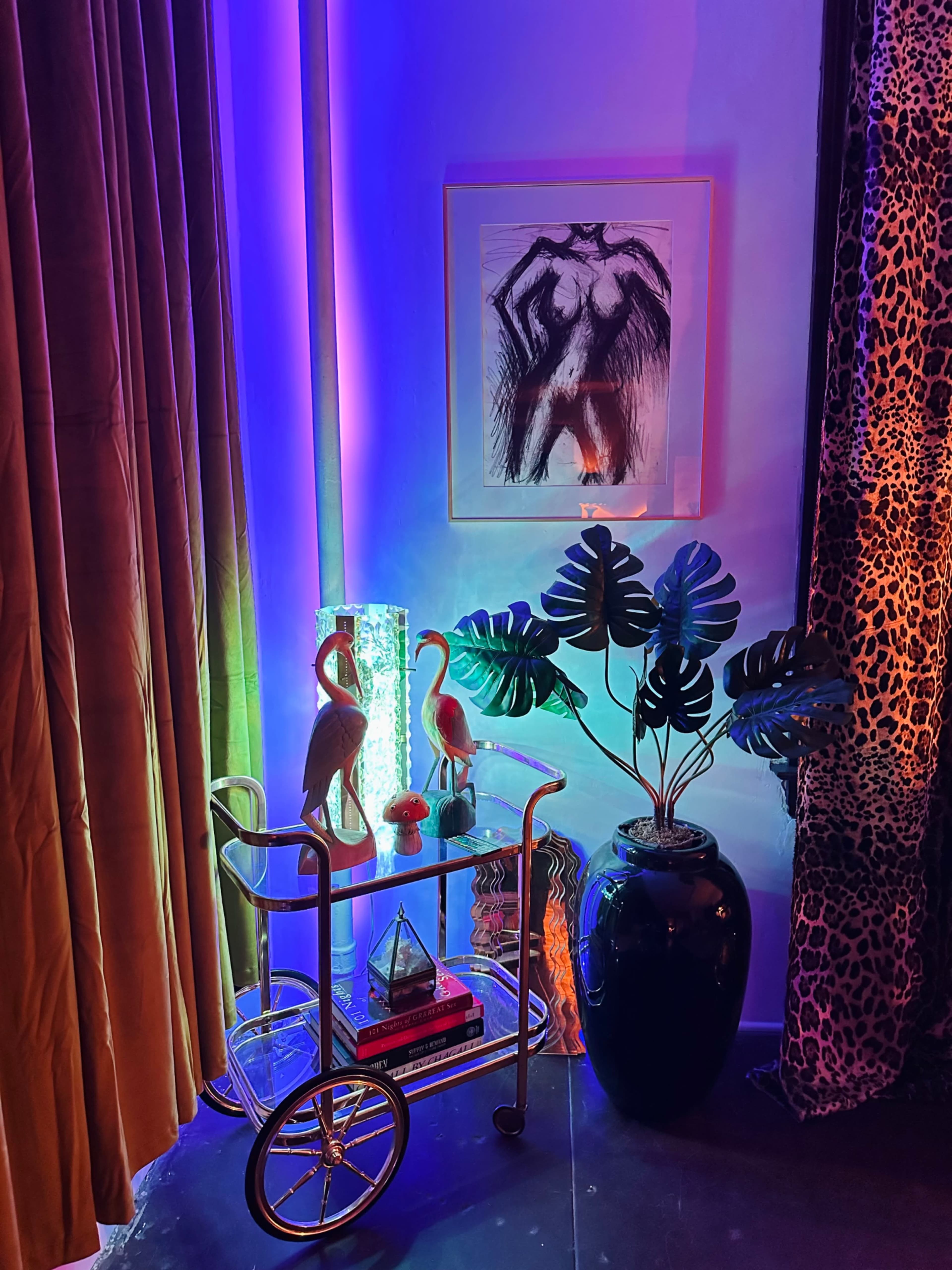 A cart with decorative items and two flamingo figurines sits beside a potted plant in a dimly lit corner with colorful lighting and patterned curtains.