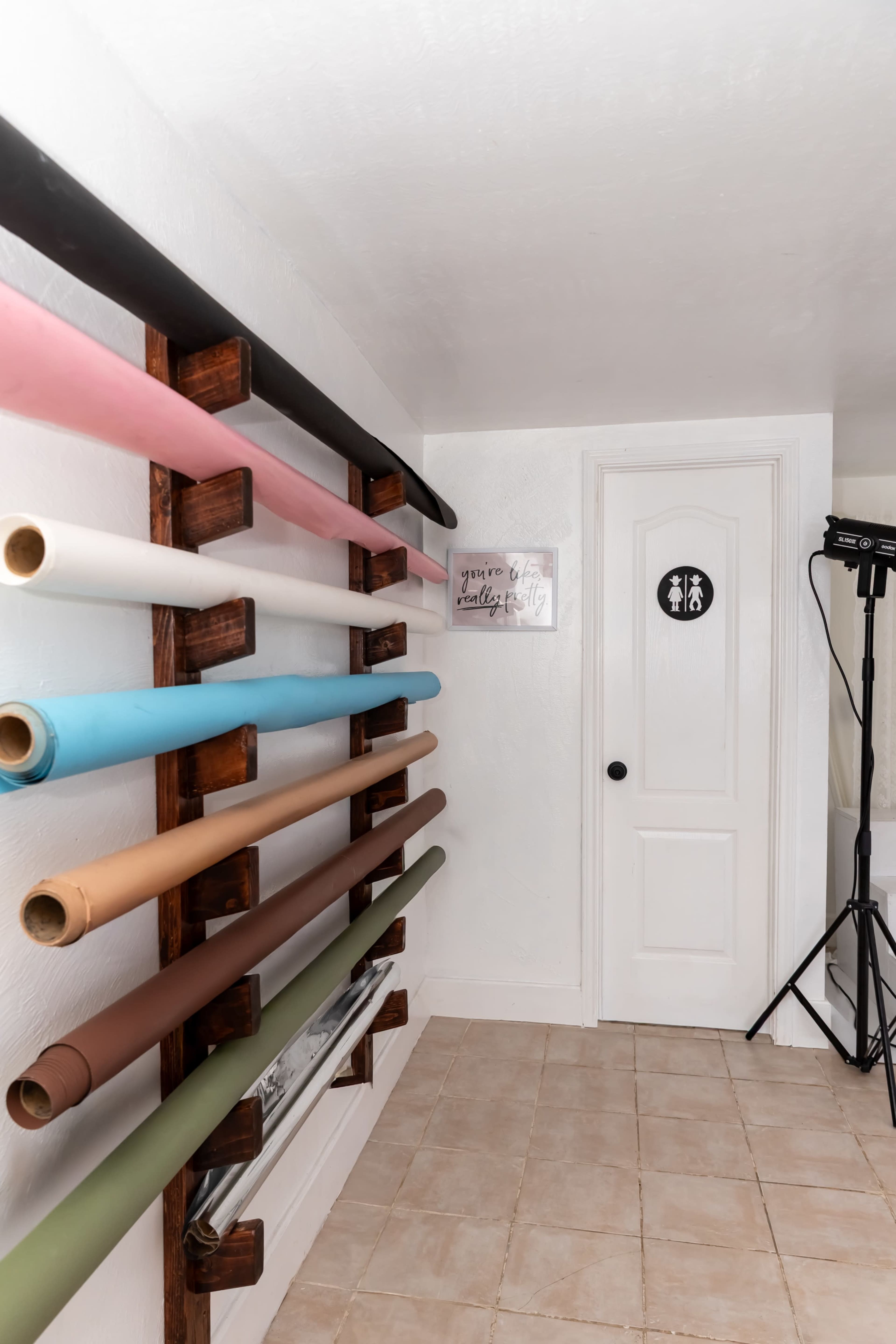 The image shows a studio space with various rolls of colored backdrops neatly organized on a wooden rack along the wall, next to a door marked with restroom signage.