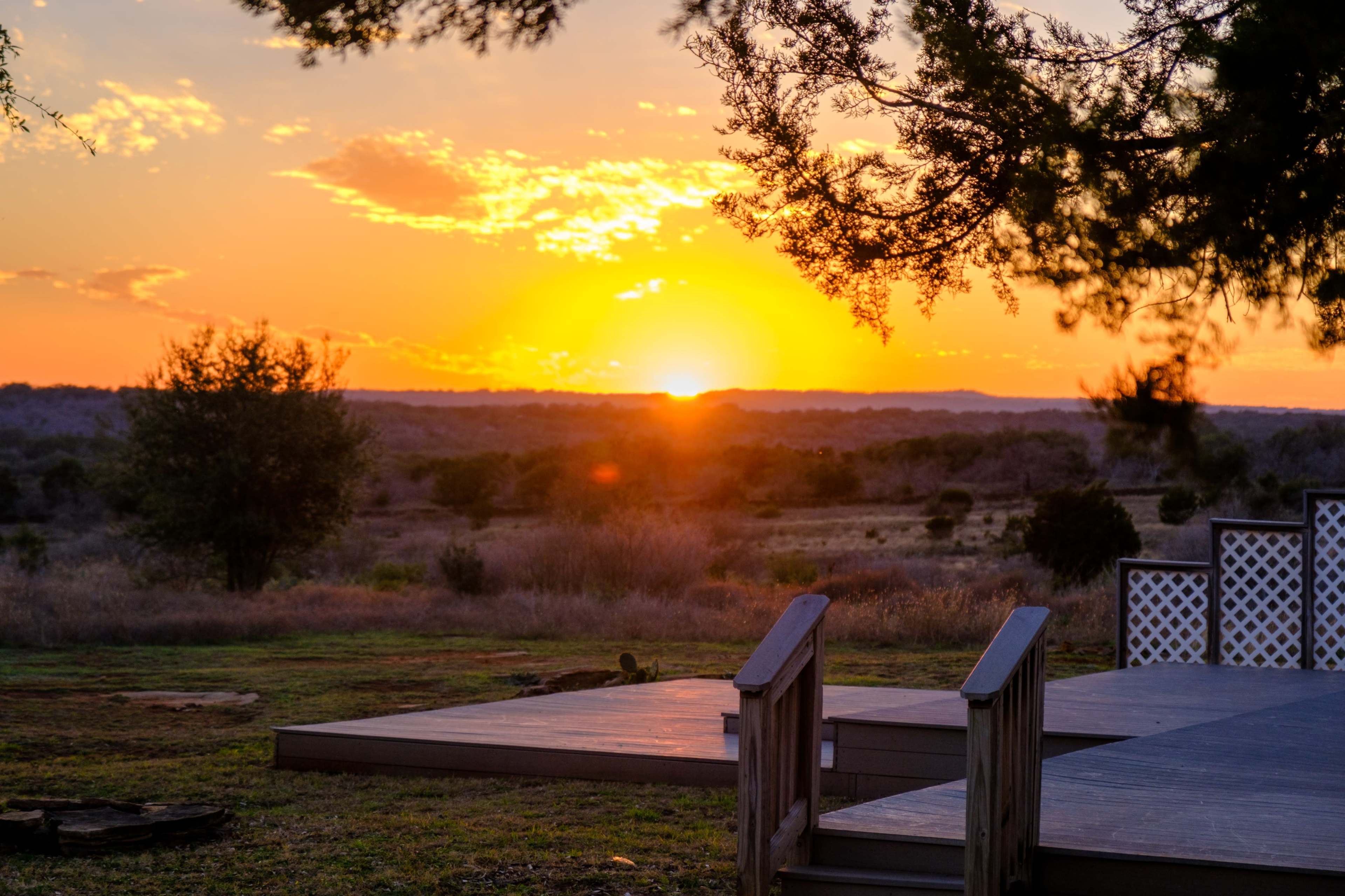 The sun sets over a landscape of open fields and distant hills, with a wooden deck in the foreground.