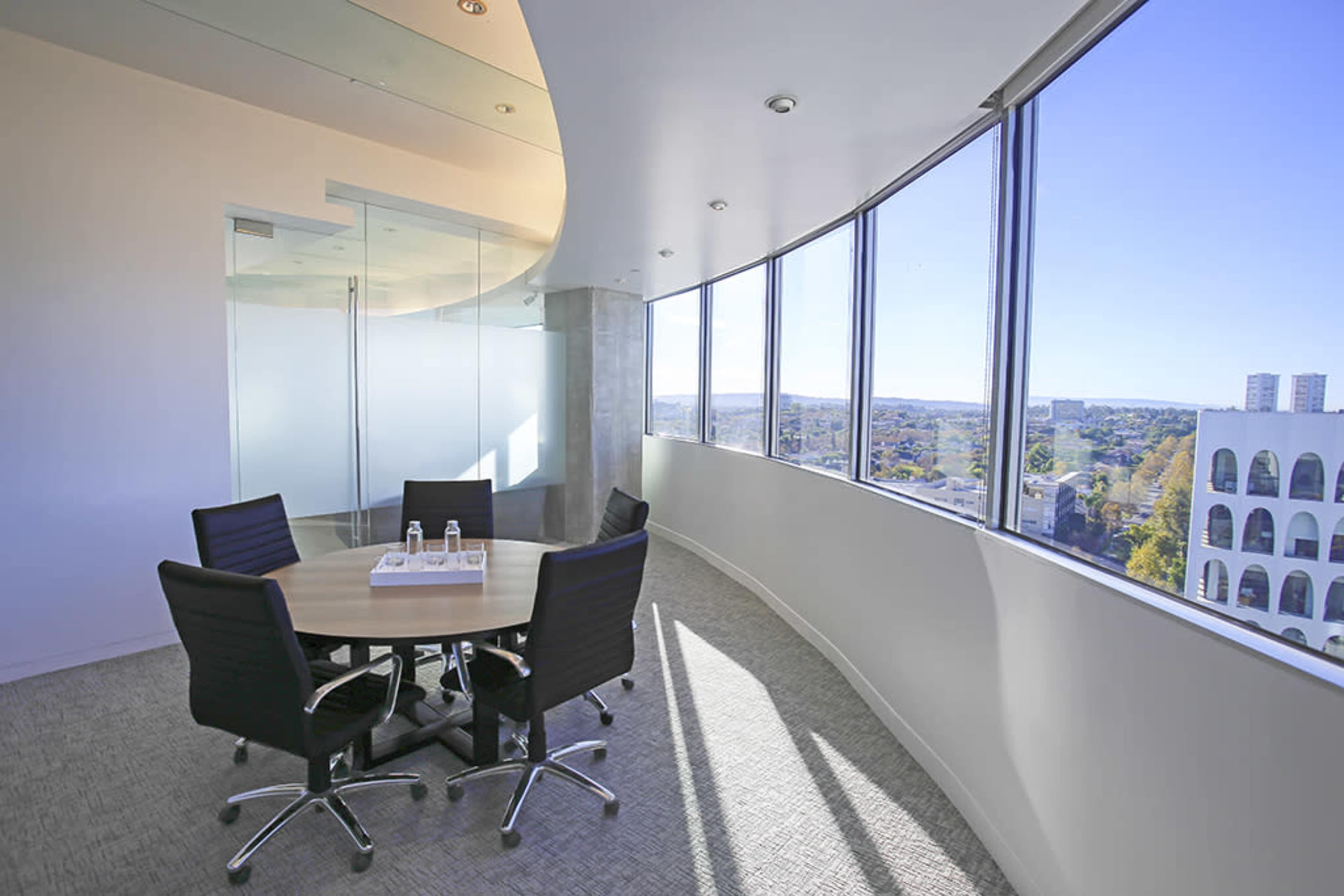 A modern conference room with a round table and four black chairs, featuring large windows that offer a view of the surrounding landscape.