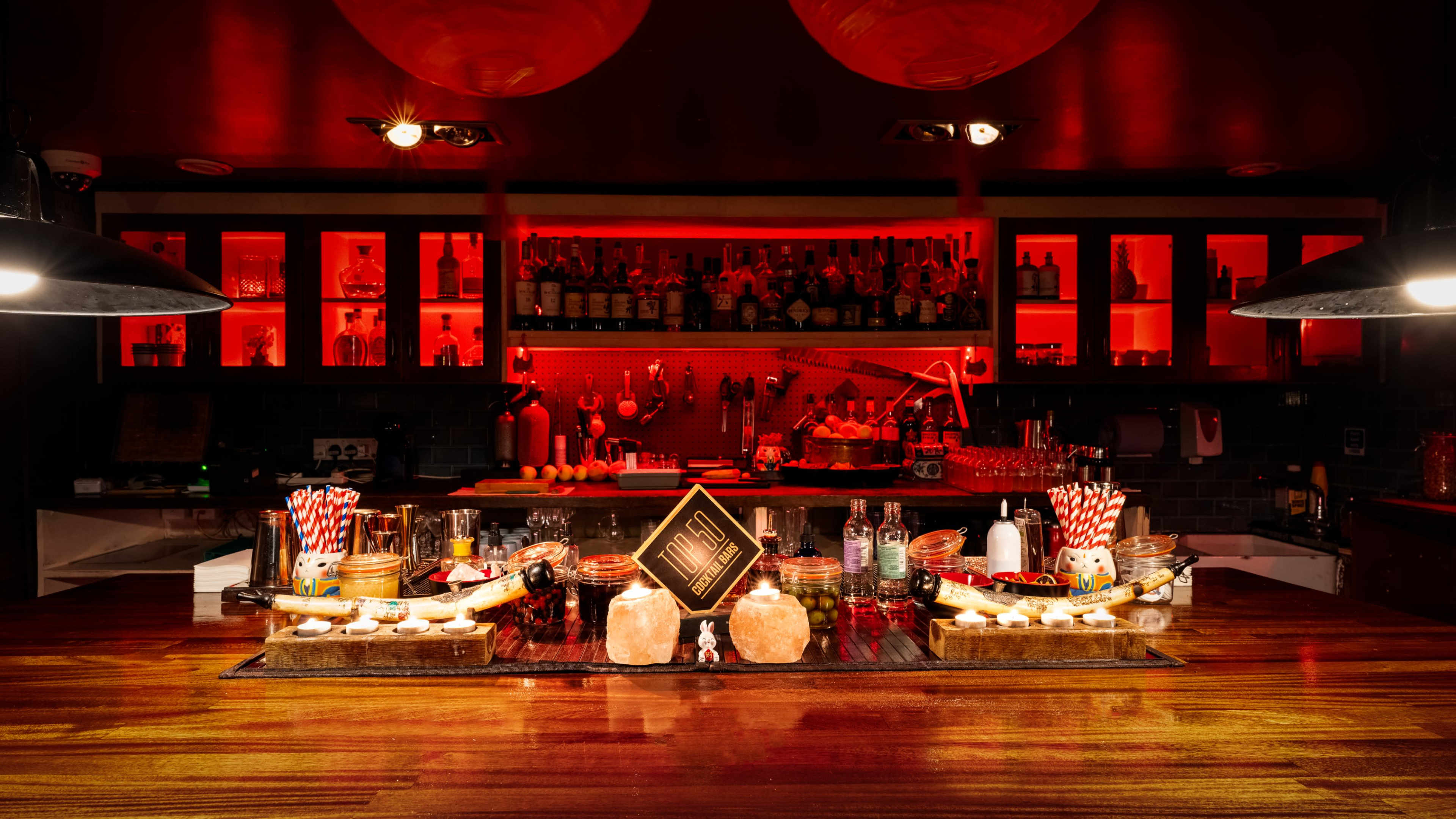 The image shows a bar counter illuminated by red lighting, featuring various bottles of liquor and an array of decorative items spread out on the surface.