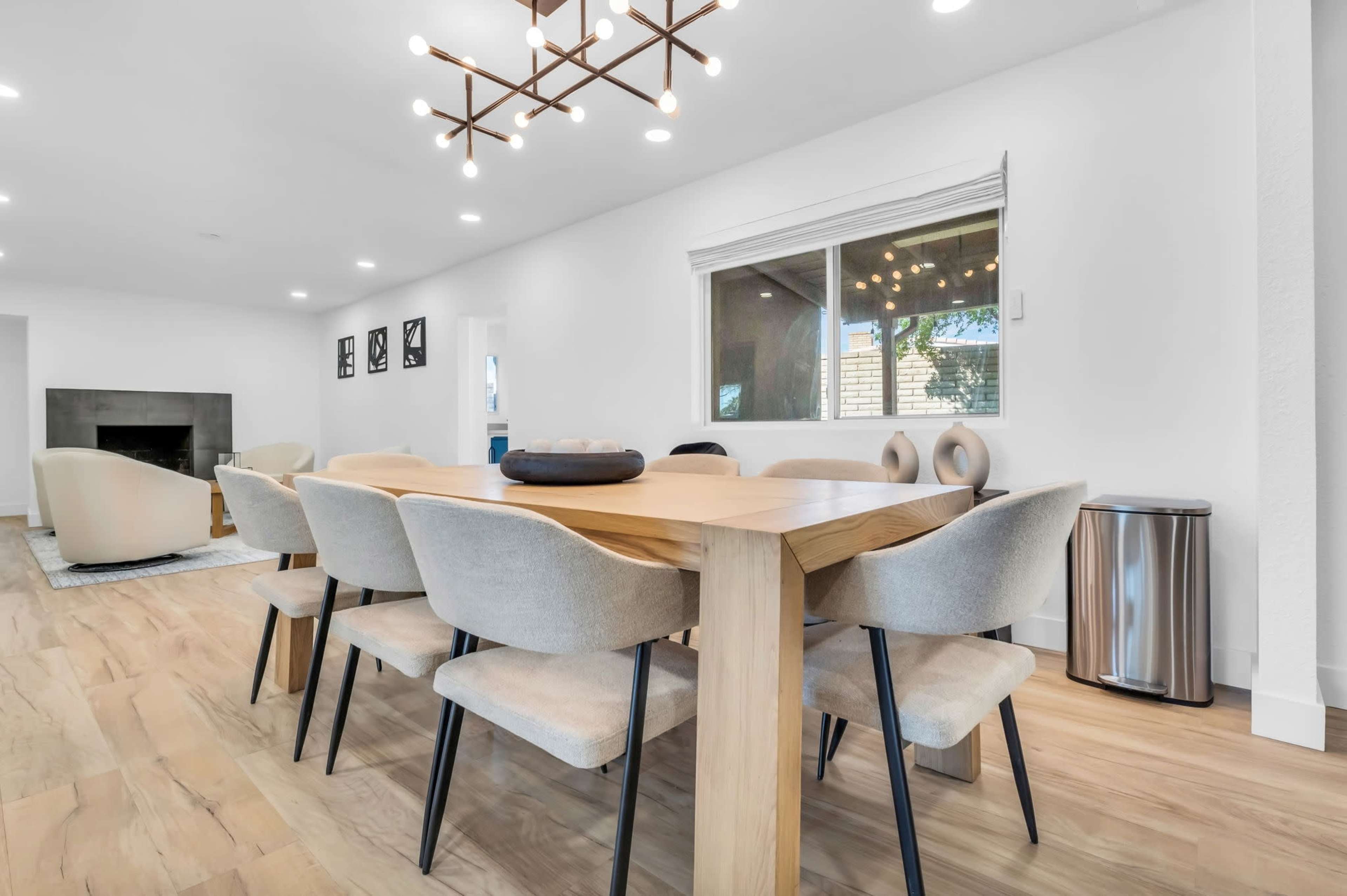 A light-filled dining area features a wooden table surrounded by upholstered chairs, with a window overlooking an outdoor space.