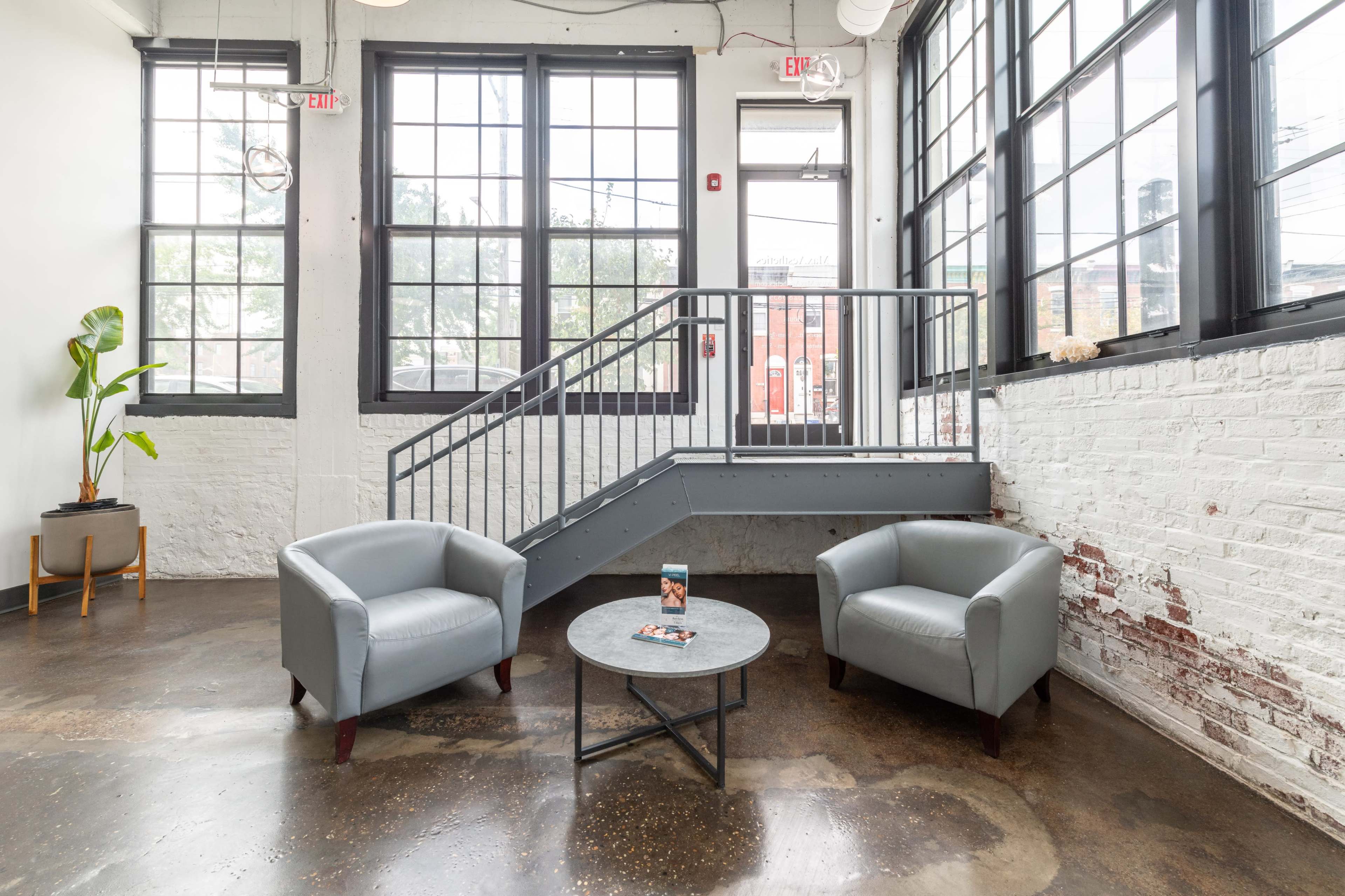 A modern interior features two gray armchairs and a circular table, with large windows allowing natural light to illuminate the space.