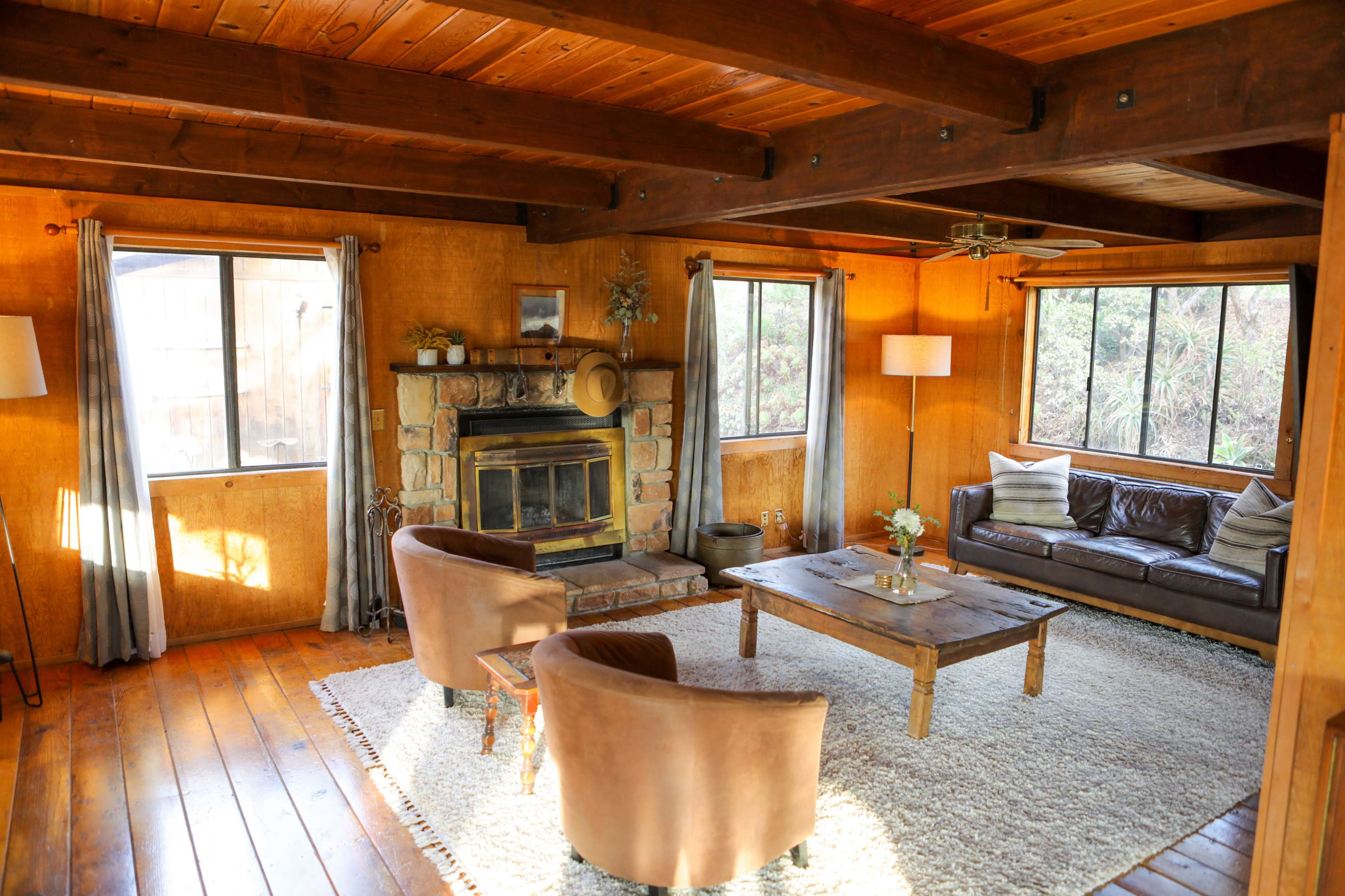 A cozy living room with wooden beams, a stone fireplace, two brown armchairs, a leather sofa, and large windows allowing natural light.