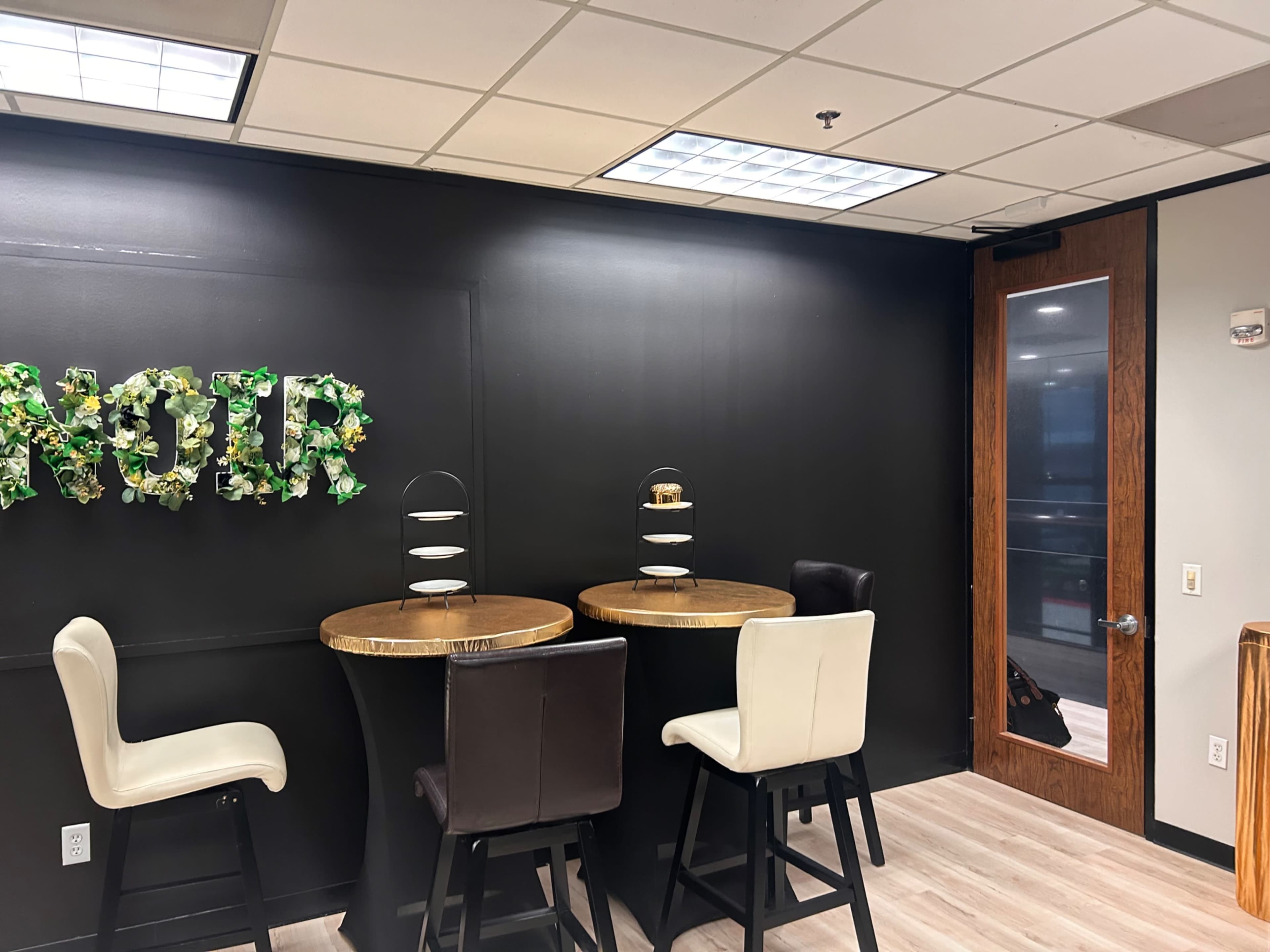 The image shows a modern office space with two round tables and high stools, against a black wall adorned with decorative text made of greenery, along with a glass door on the right.