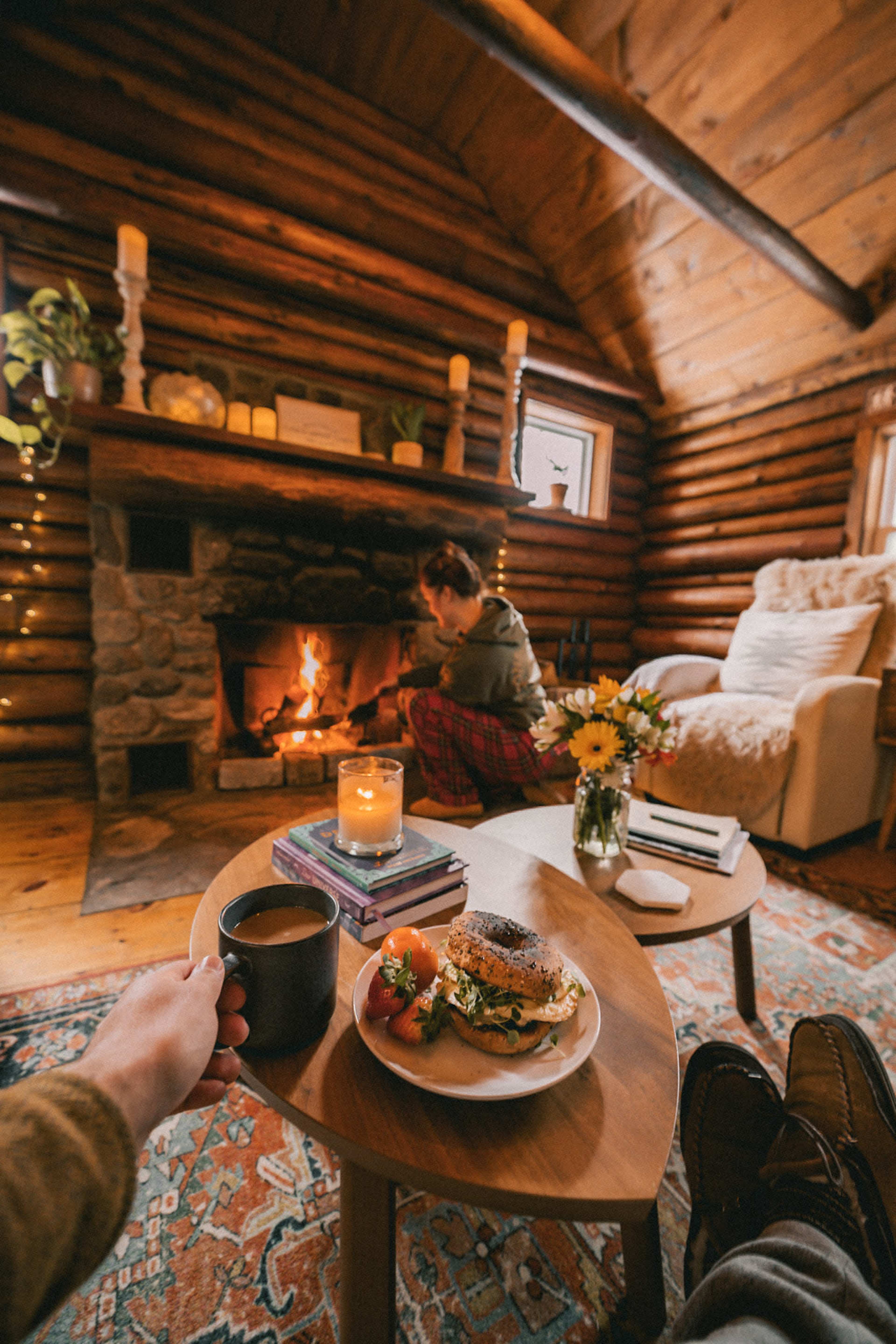 A person sits in a cozy cabin with a fire burning in the stone fireplace, while holding a plate of food and a cup of coffee on a small table.