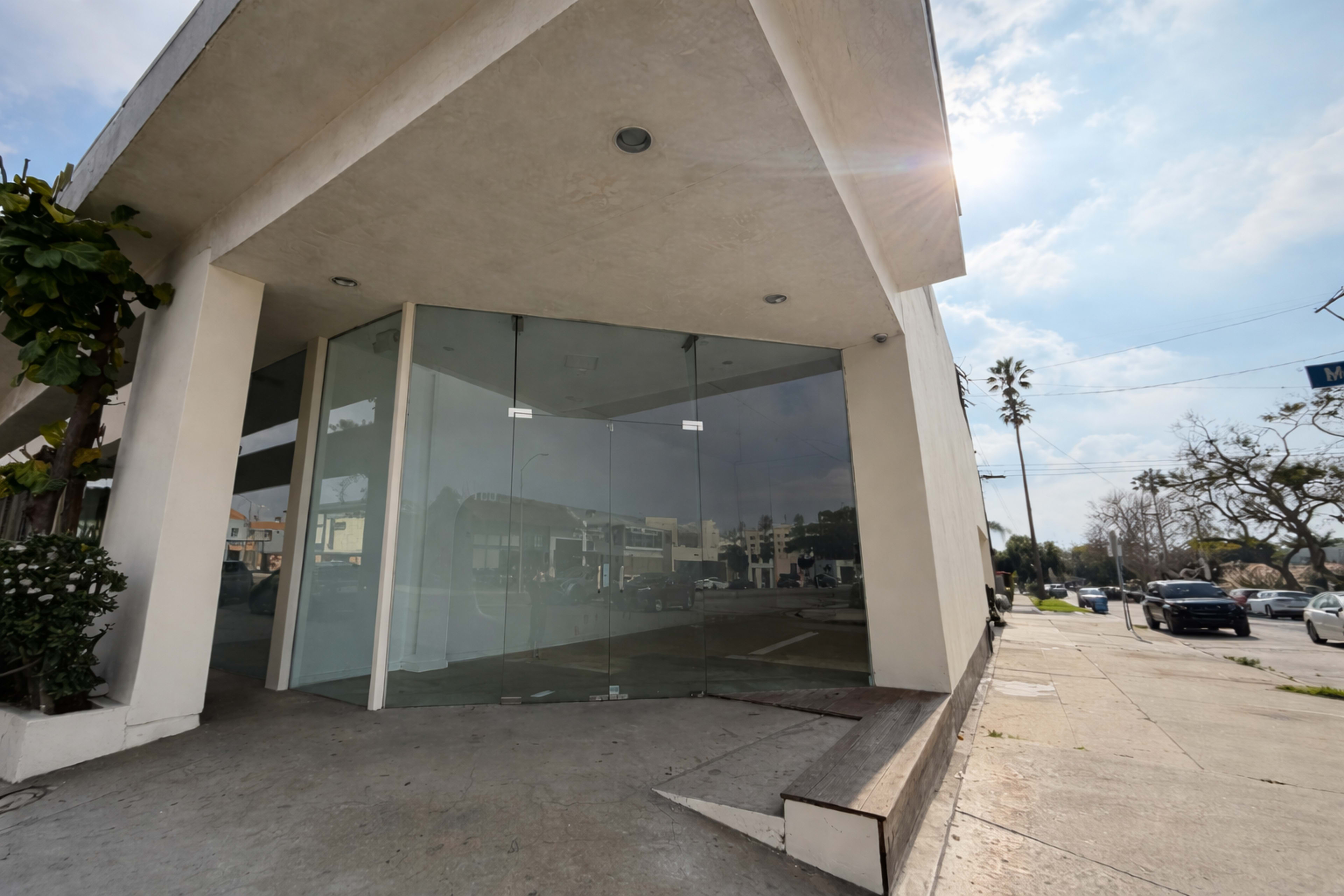 A vacant commercial building with large glass windows is situated on a street corner, surrounded by greenery and parked cars.