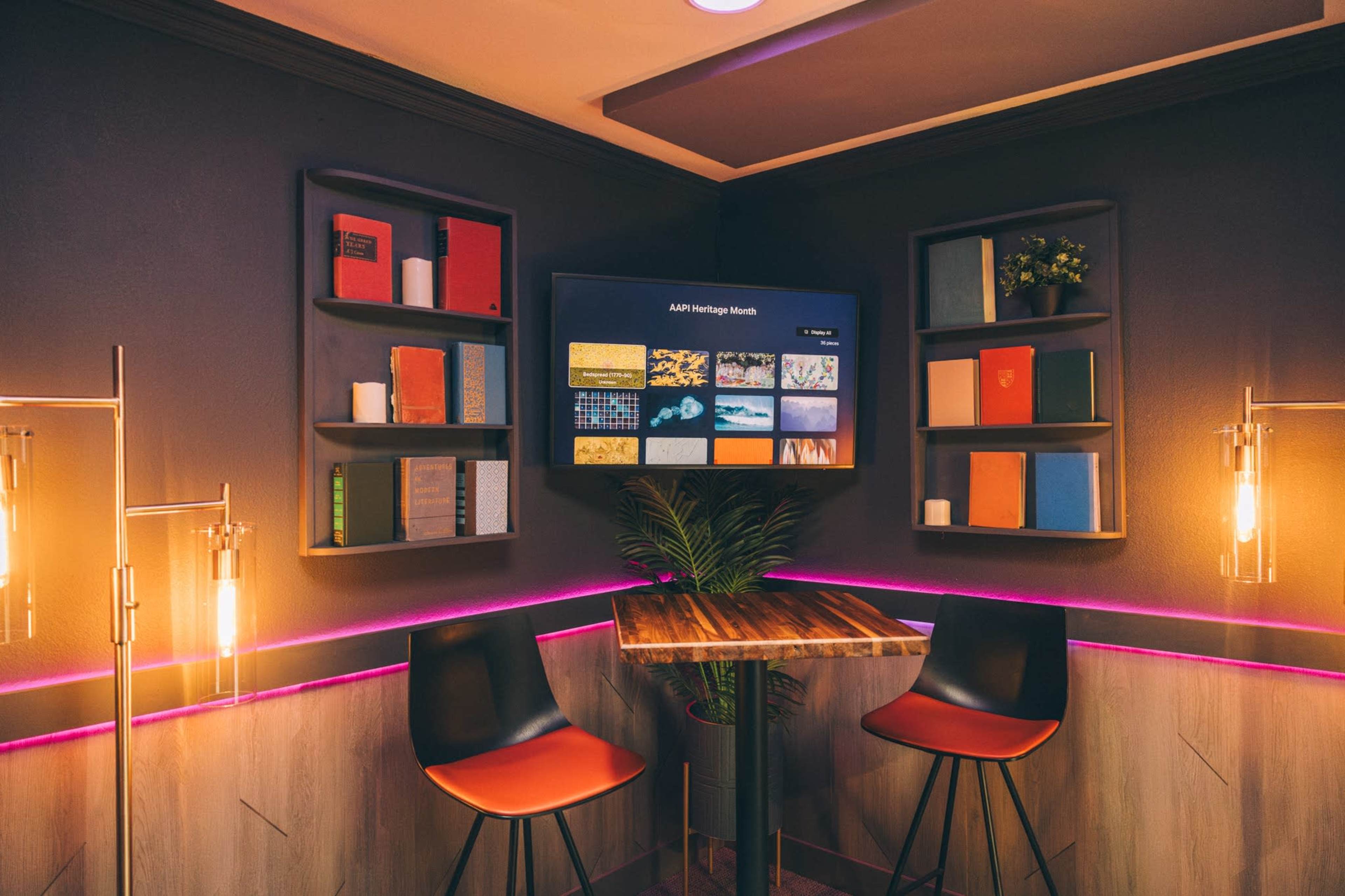 A modern corner workspace features two black chairs around a small table, flanked by shelves displaying books and a wall-mounted screen.