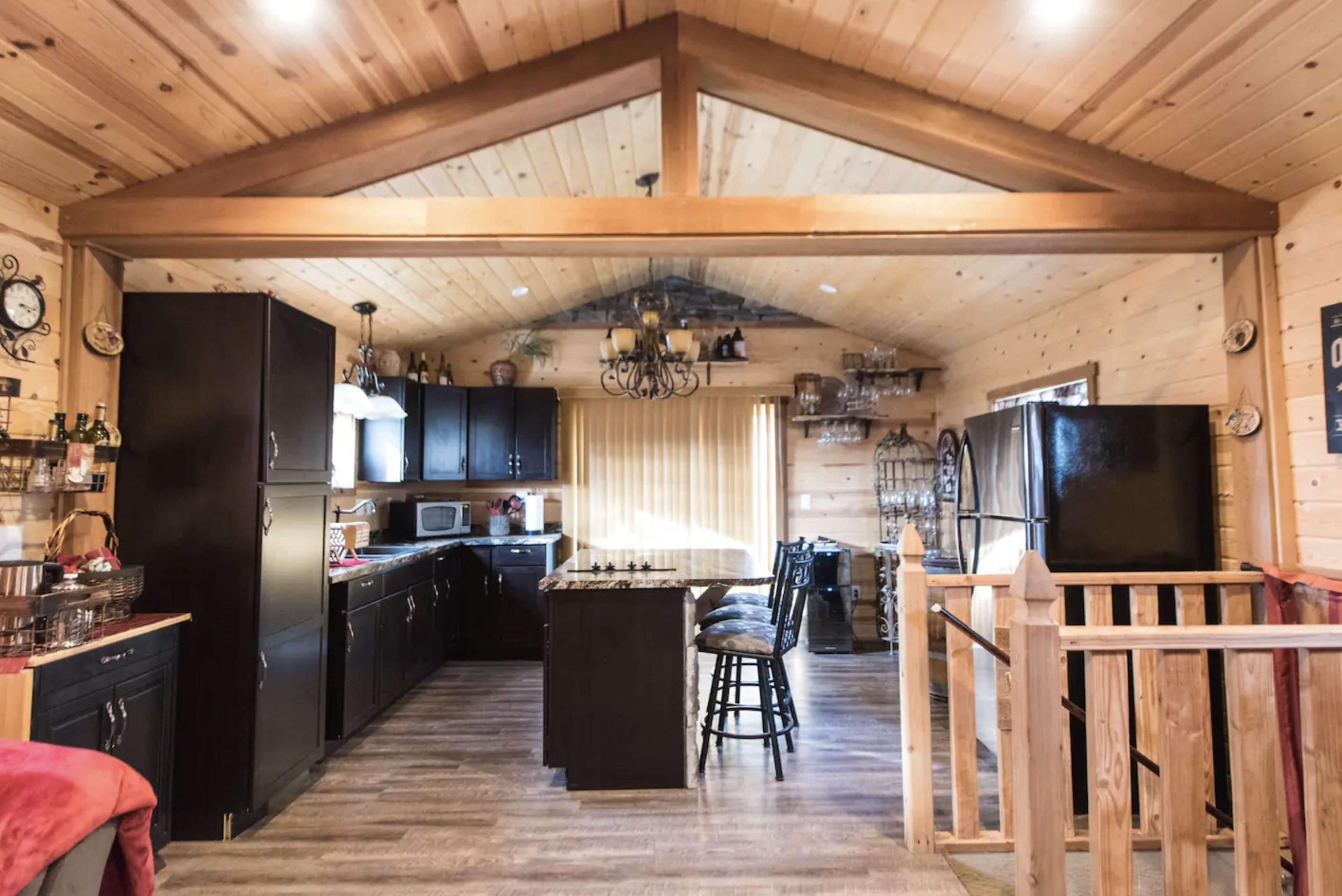 The image shows a wooden cabin kitchen with black cabinets, a central island, and a spacious dining area featuring a large window.