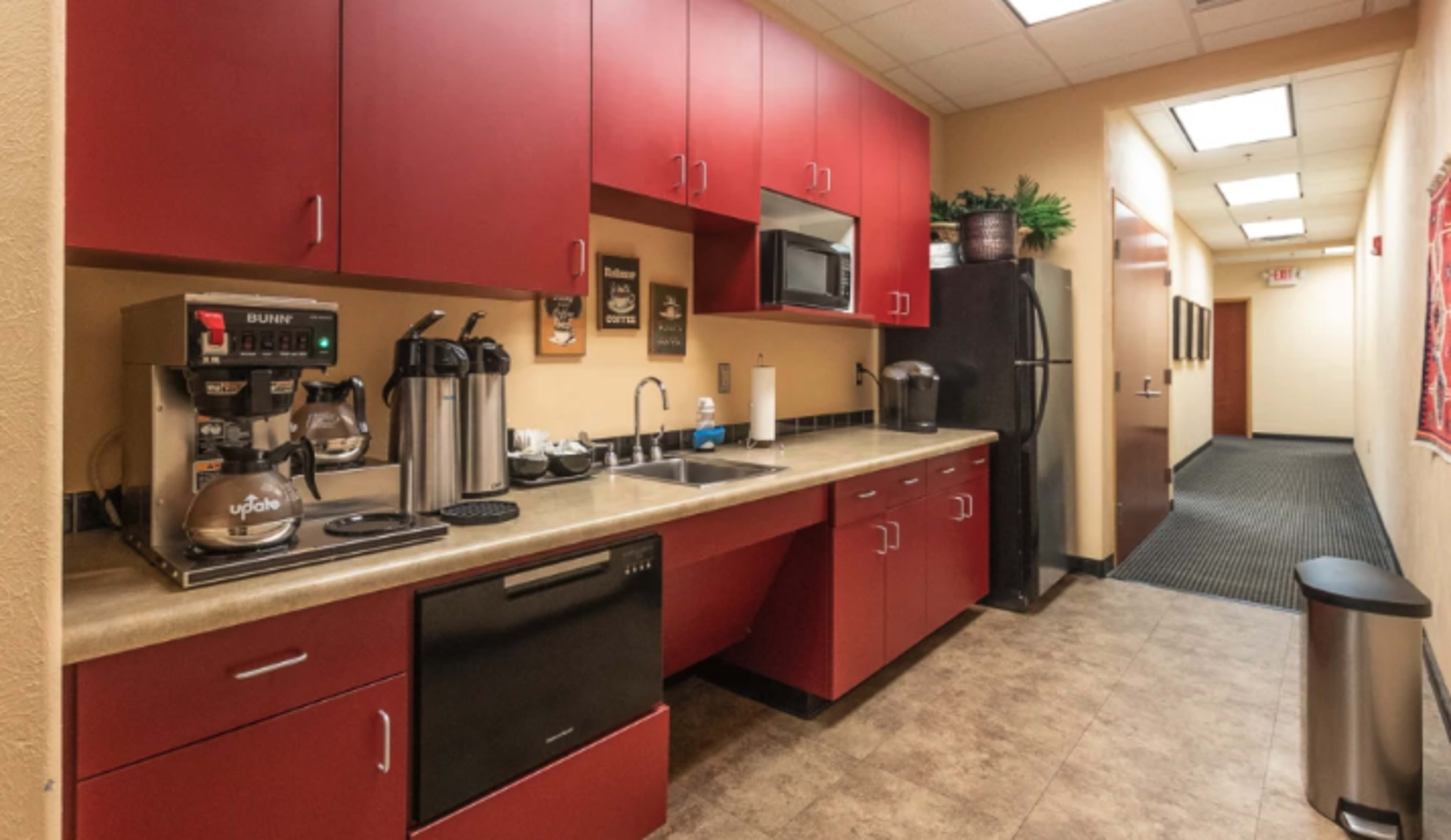 A kitchen area featuring red cabinets, a coffee machine, microwave, and a black refrigerator, adjacent to a hallway.