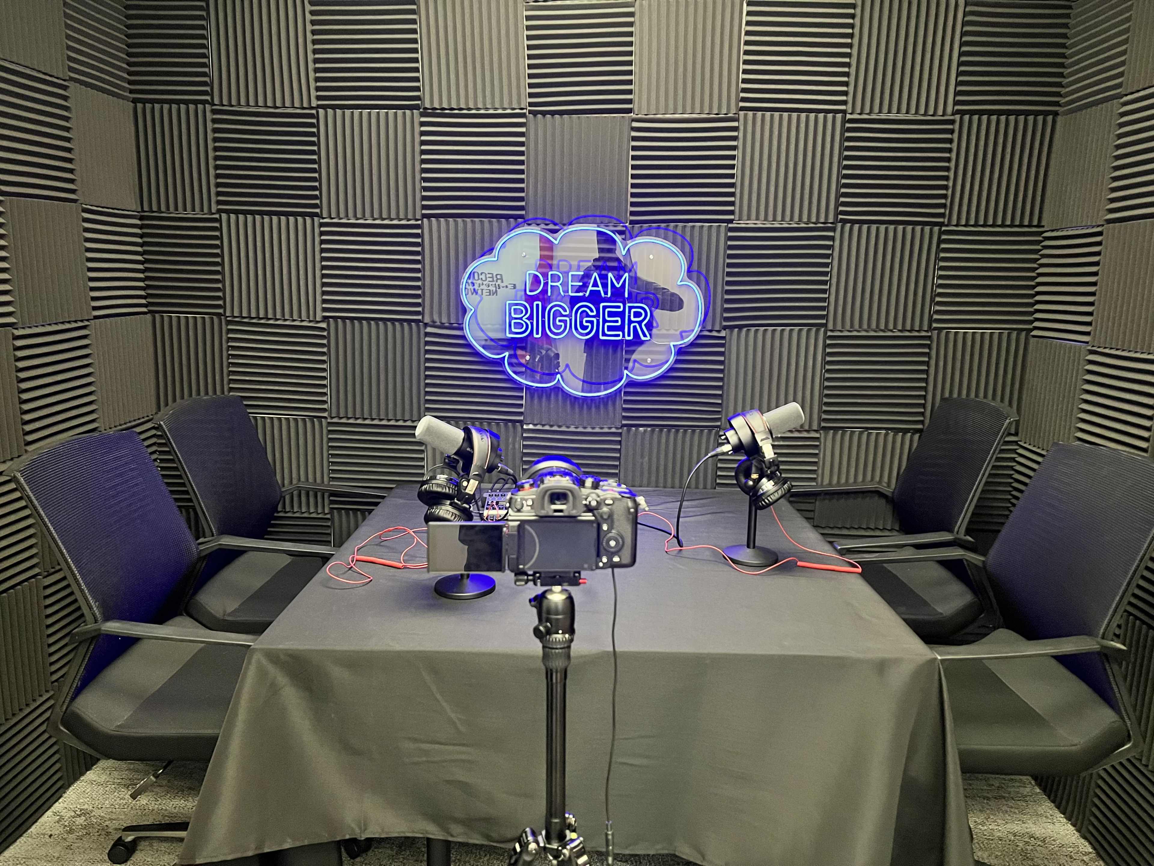 A podcast recording studio features a black table, four chairs, two microphones on stands, and a camera set up, with a neon sign that says "DREAM BIGGER" on the wall.