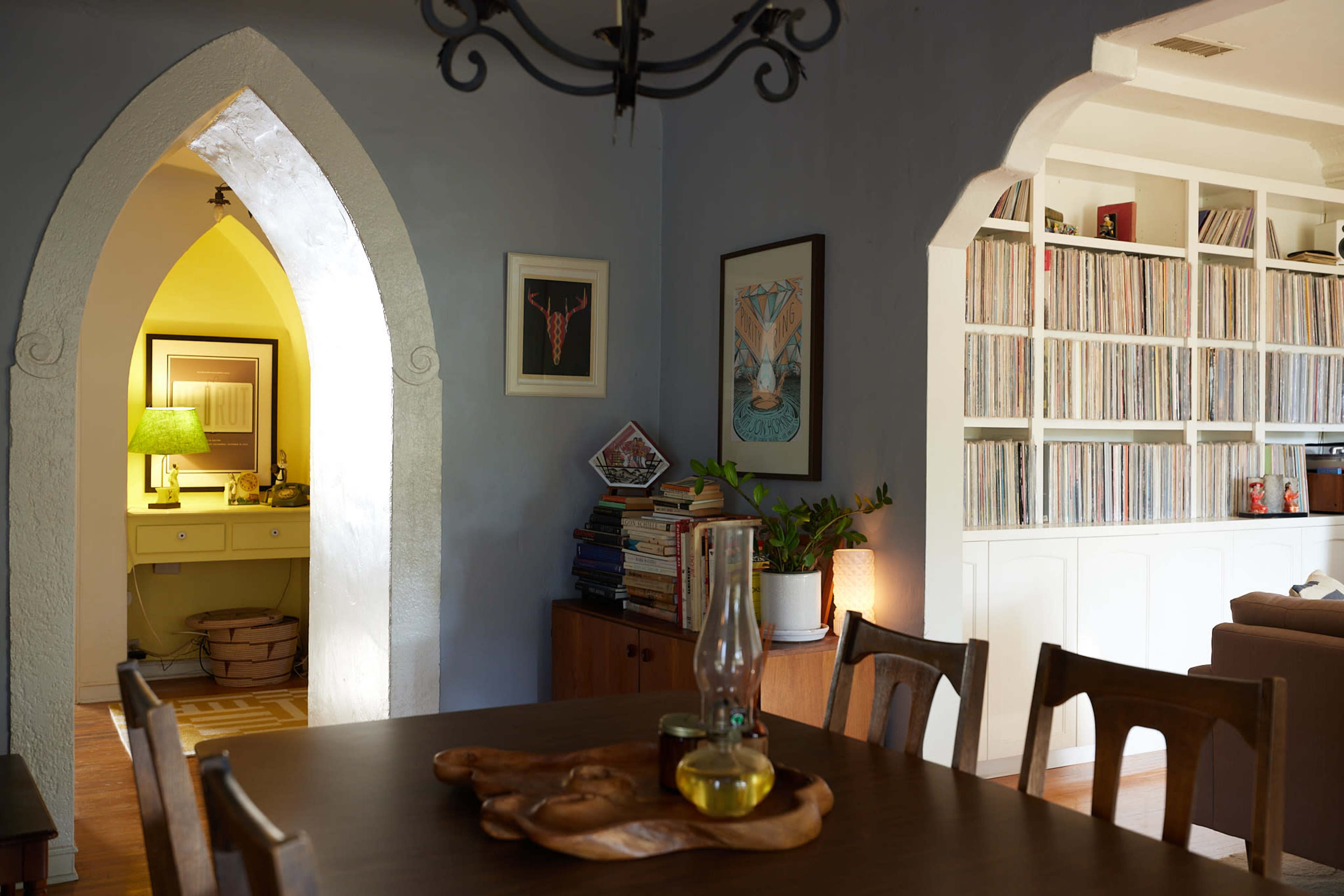 The image shows a dining area with a wooden table, an oil lamp, and arched doorways leading to a room with a desk and a bookshelf filled with records.