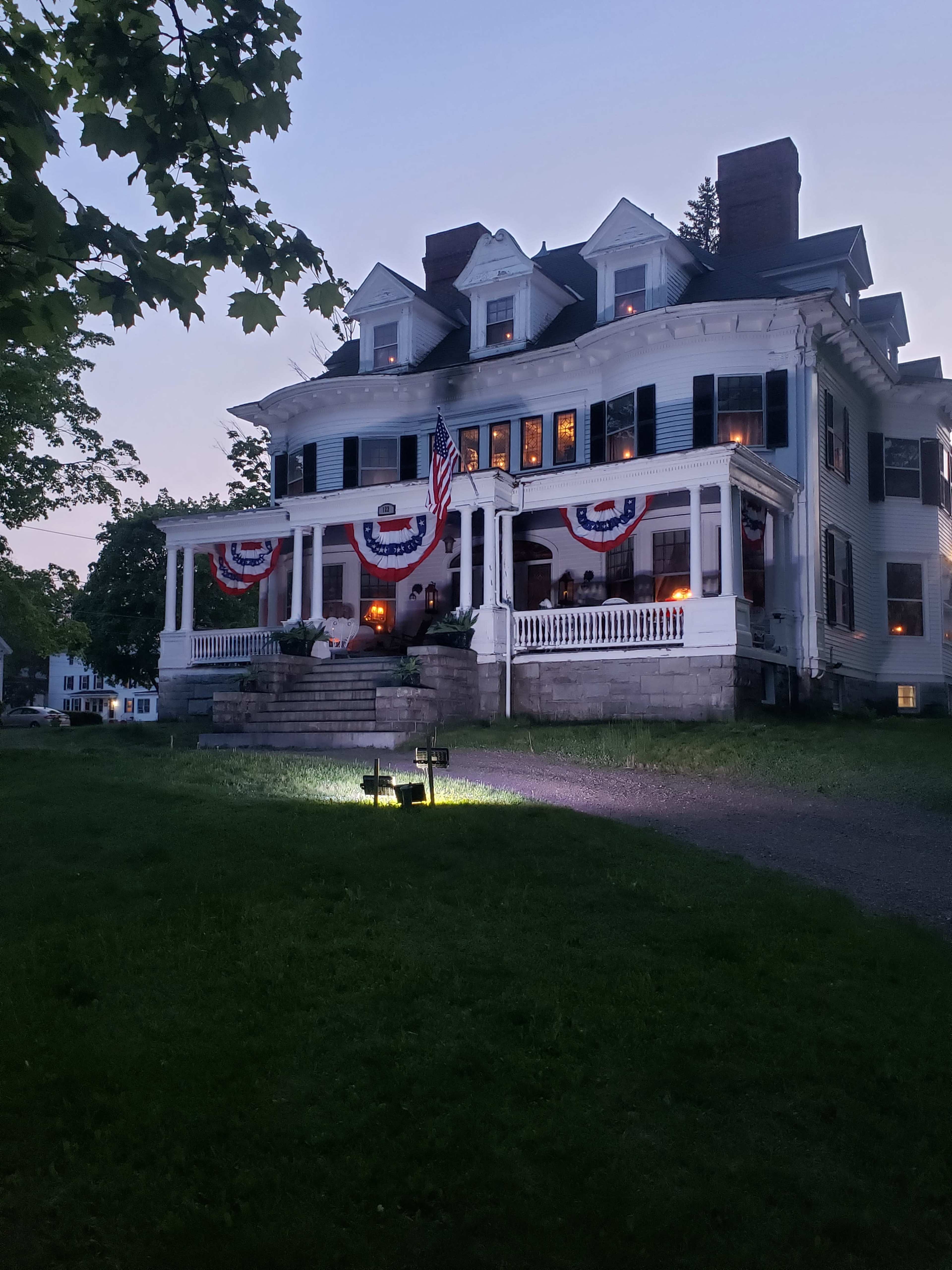 A large, historic house is adorned with patriotic bunting and illuminated by outdoor lighting during twilight.
