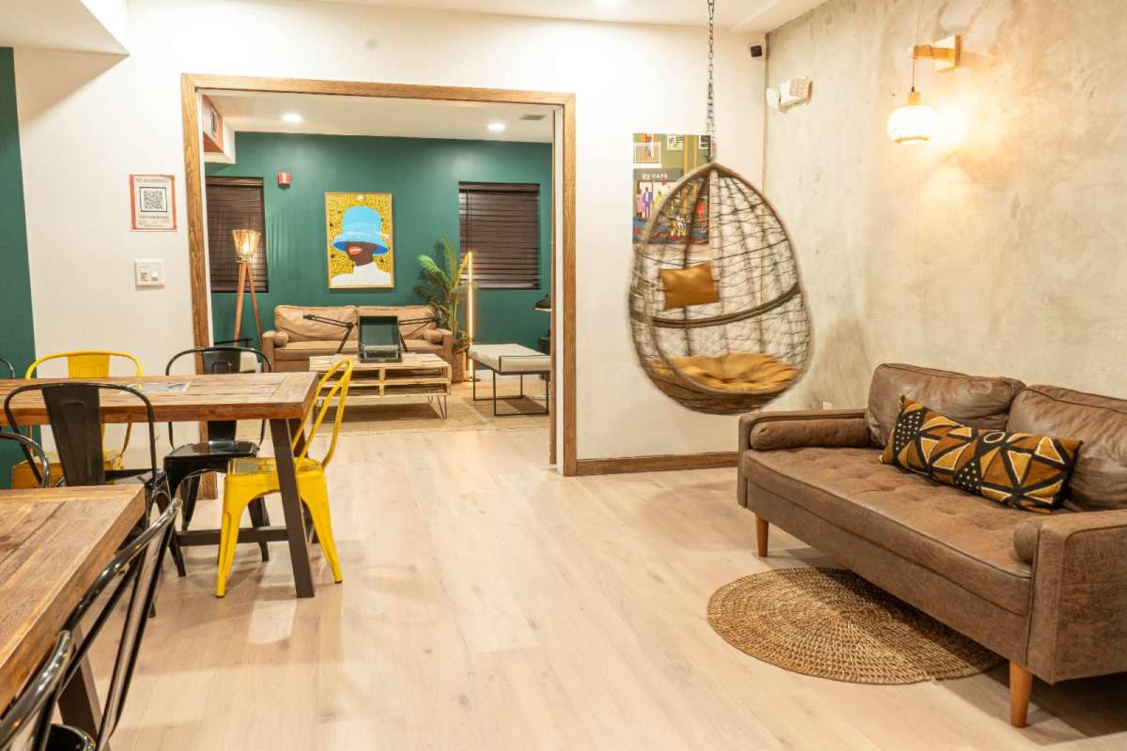 The image shows a modern, well-lit lounge area featuring a hanging rattan chair, a brown sofa, and a dining table with colorful chairs.