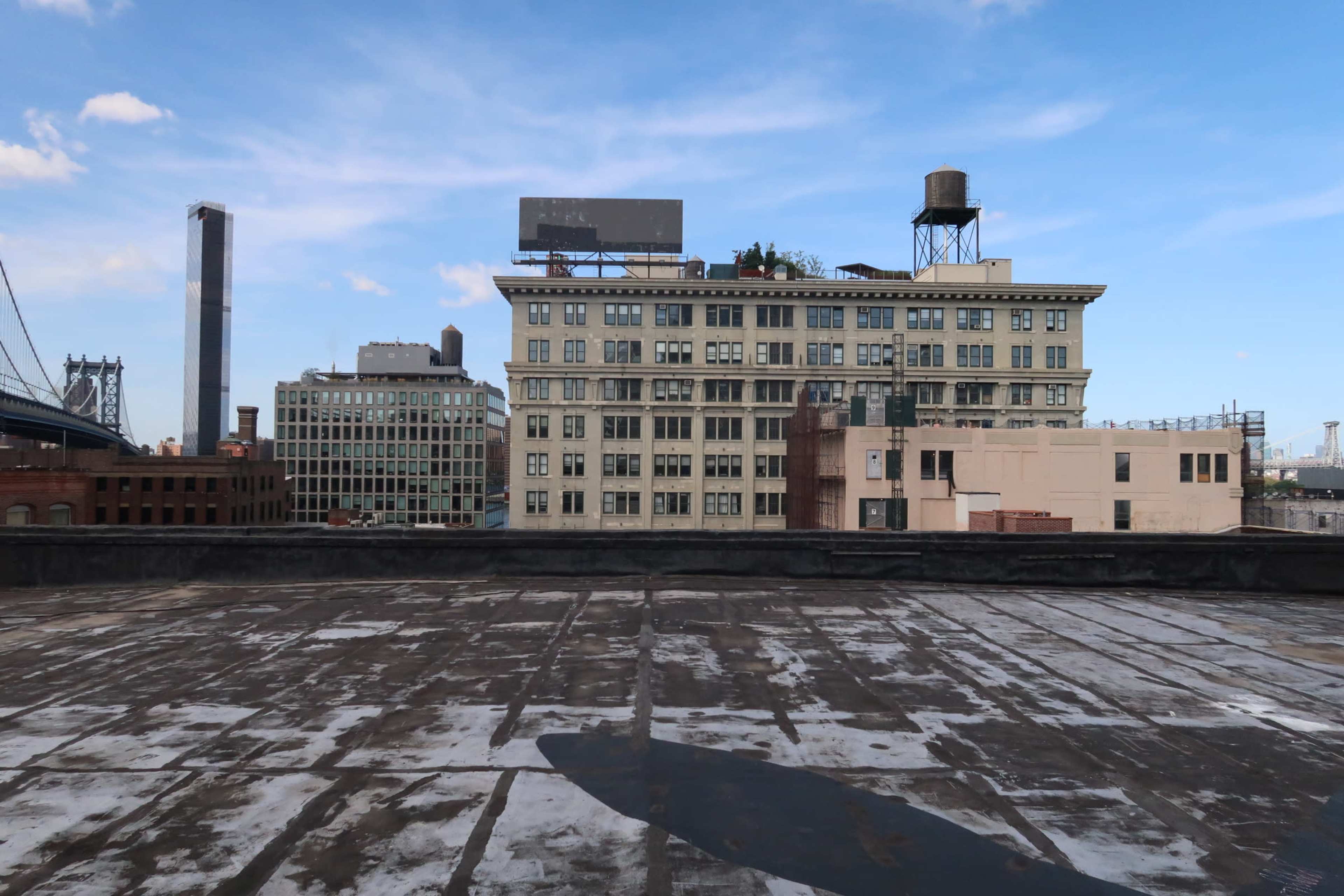 A rooftop view of buildings, including a water tower and billboards, against a blue sky with scattered clouds.