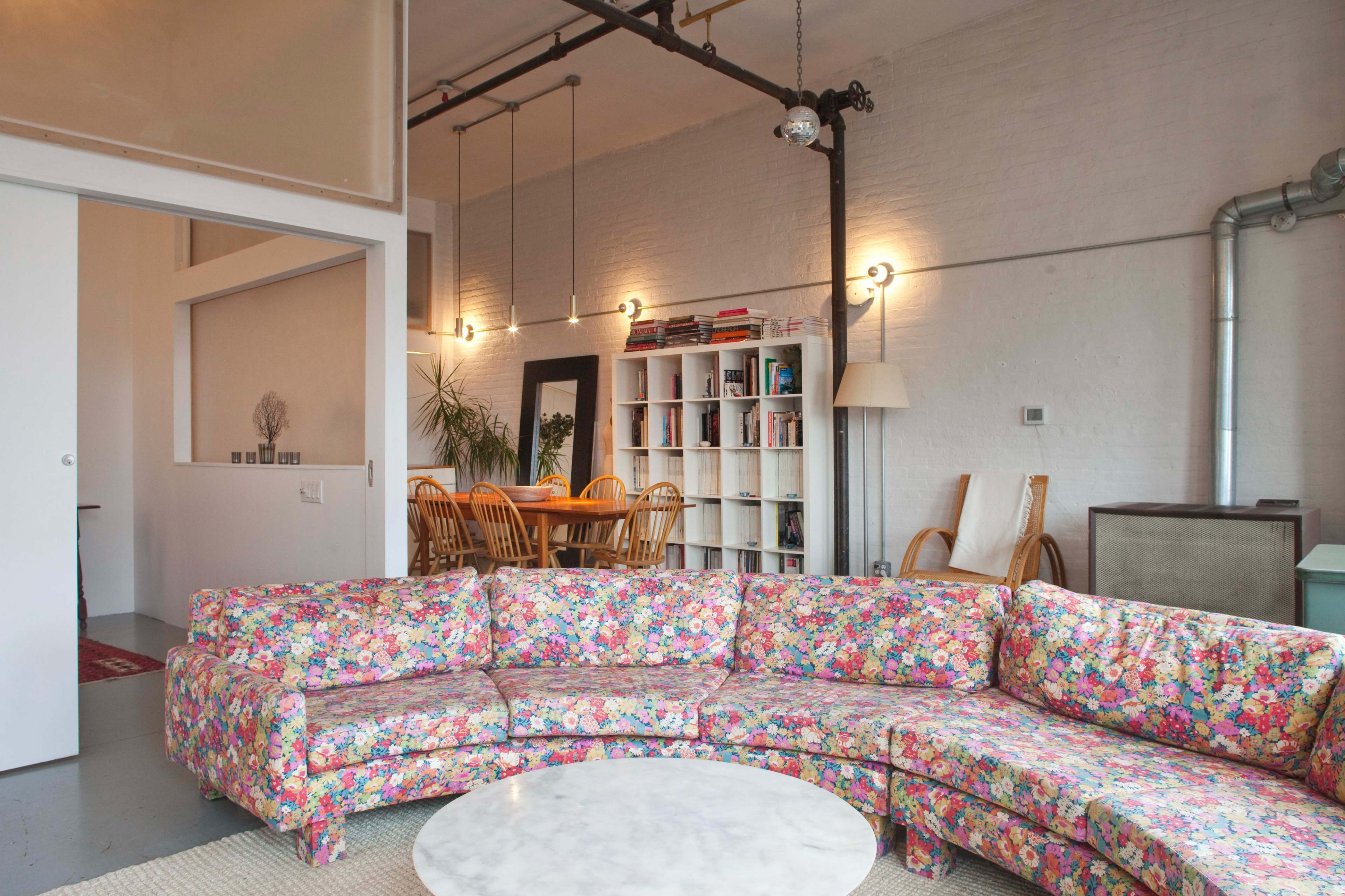 A colorful floral-patterned sectional sofa surrounds a round marble coffee table in a modern, open-plan living space that connects to a dining area.