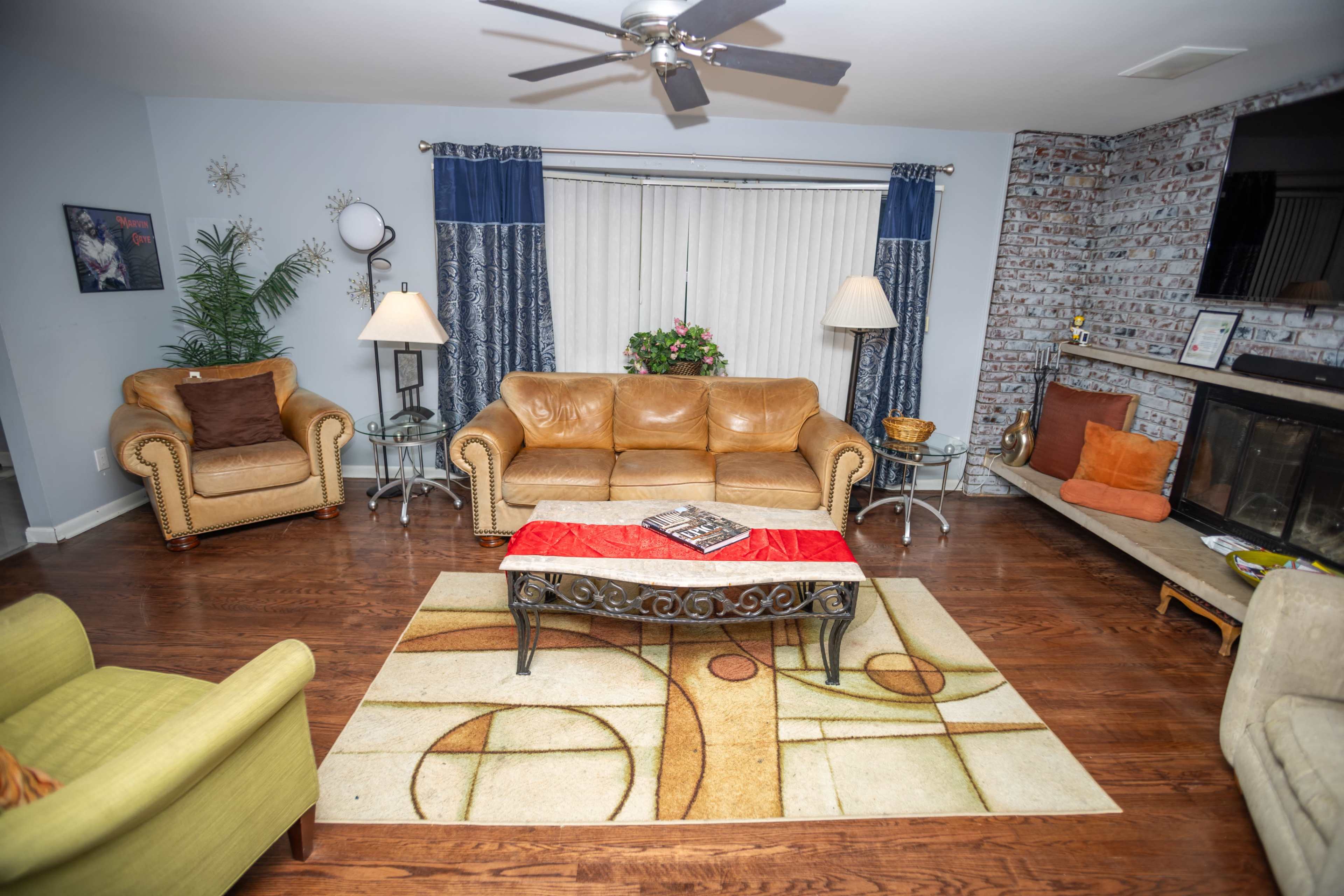 The living room features a mix of seating, including tan leather sofas and a green chair, arranged around a coffee table on a patterned area rug, with a fireplace and decorative elements in the background.