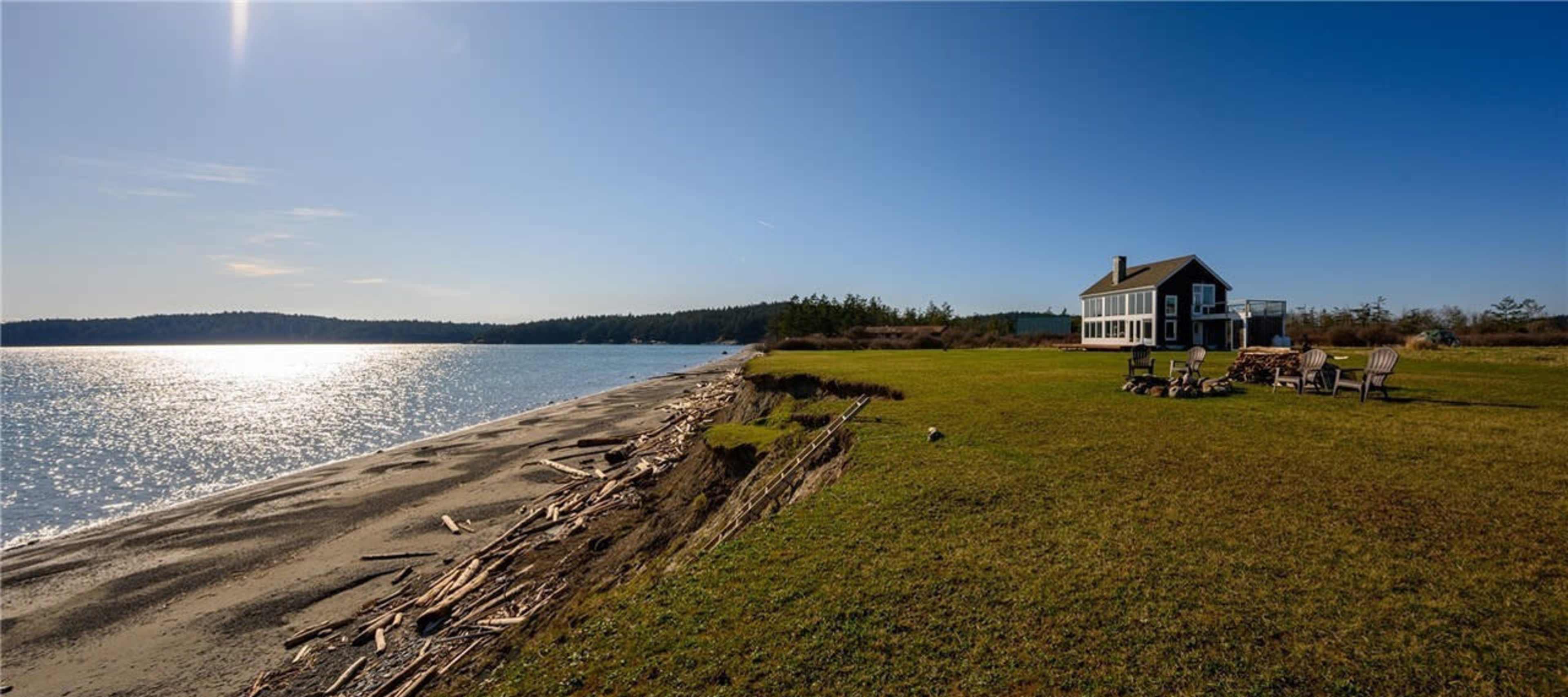 A beach house sits on a grassy shore, with driftwood scattered along the water's edge and a clear blue sky overhead.