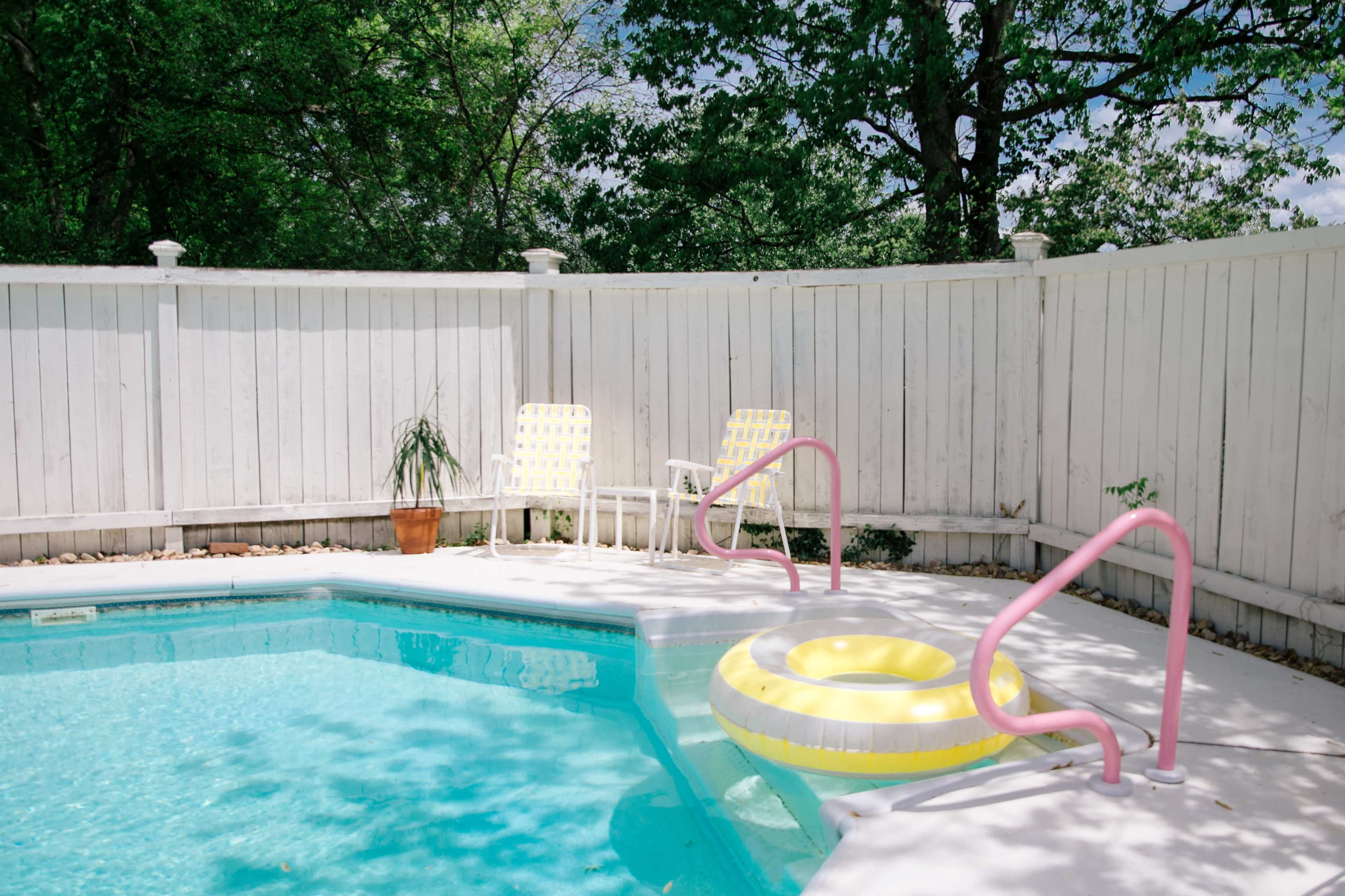 A swimming pool area features a yellow float, white patio chairs, and pink railing surrounded by trees and a white fence.