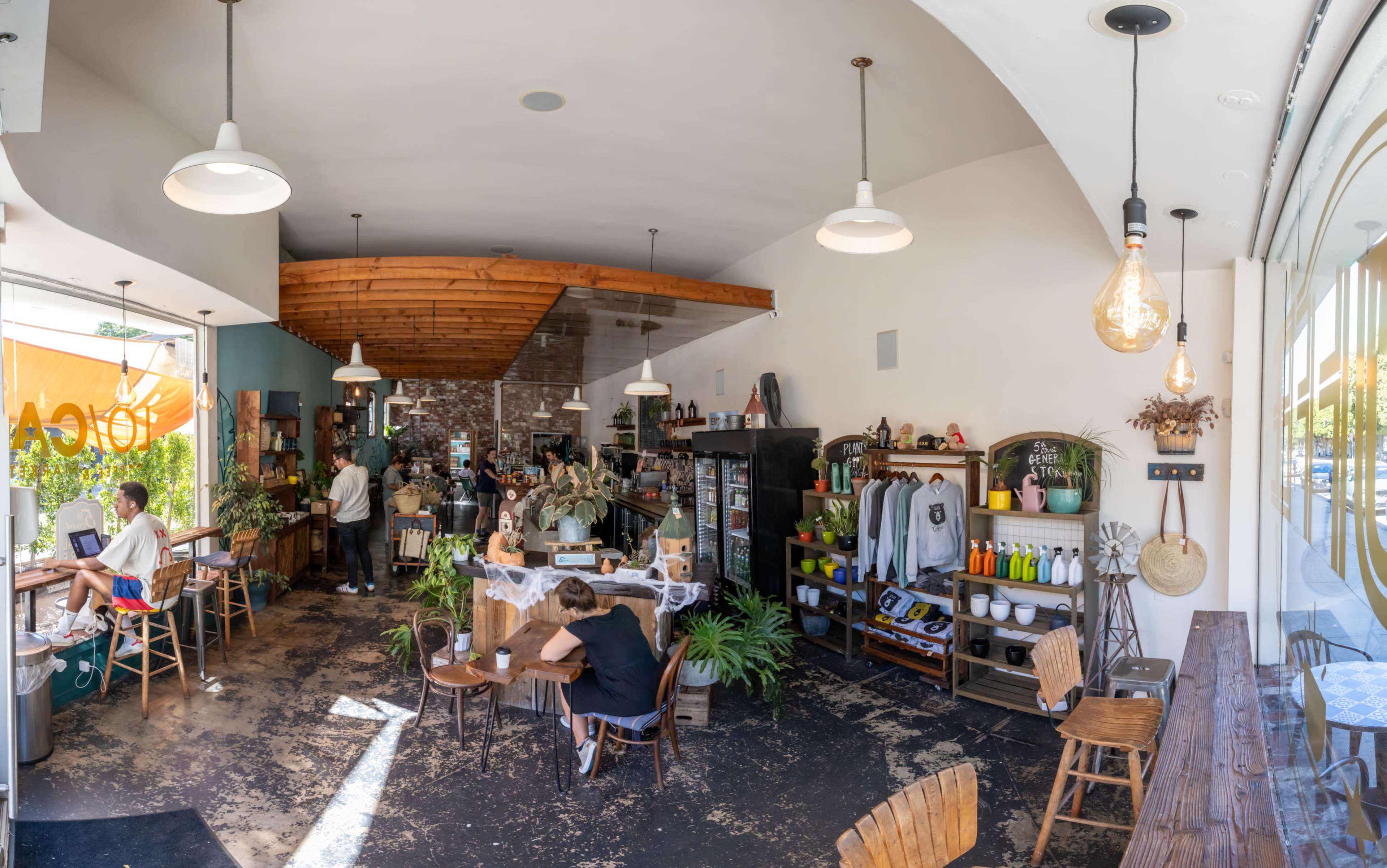 A spacious café interior with wooden furniture, plants, and customers engaging in various activities.
