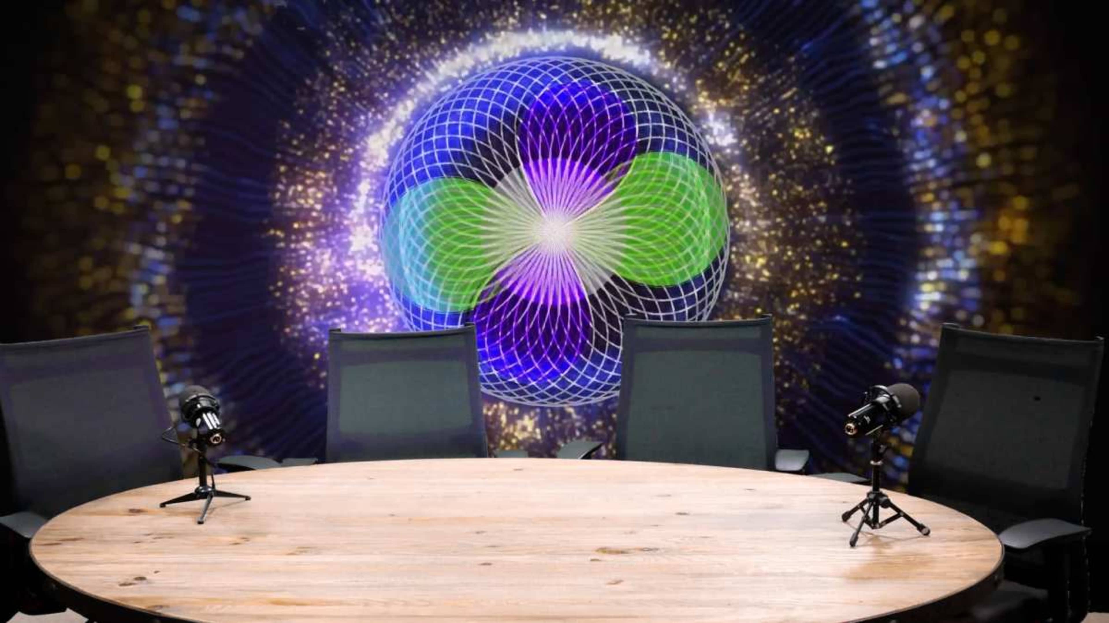 A round wooden table is set in front of several black chairs, with microphones positioned on either side and a colorful digital backdrop featuring a geometric design.
