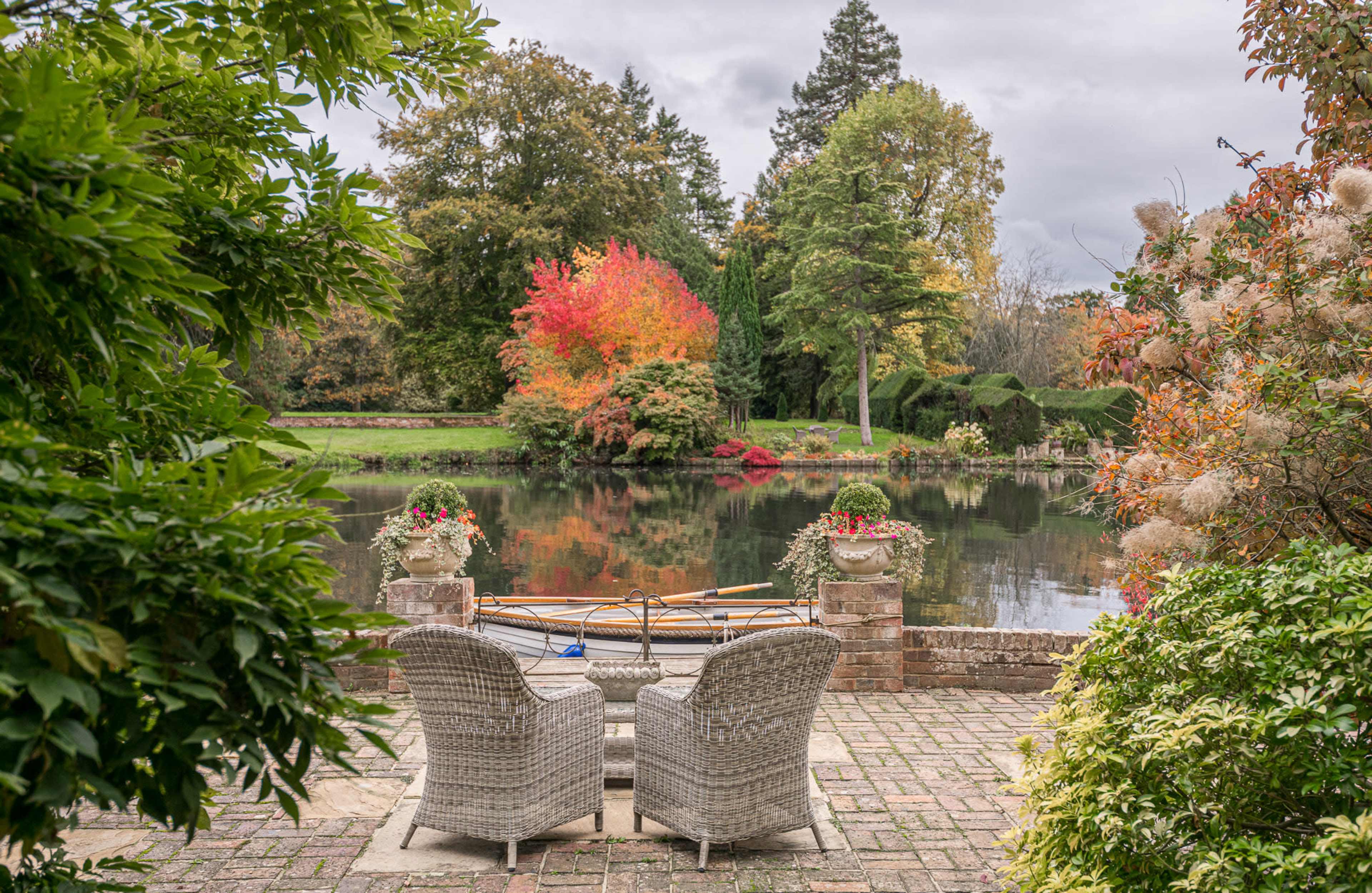 A pair of wicker chairs sits on a brick patio overlooking a tranquil lake surrounded by colorful autumn foliage.