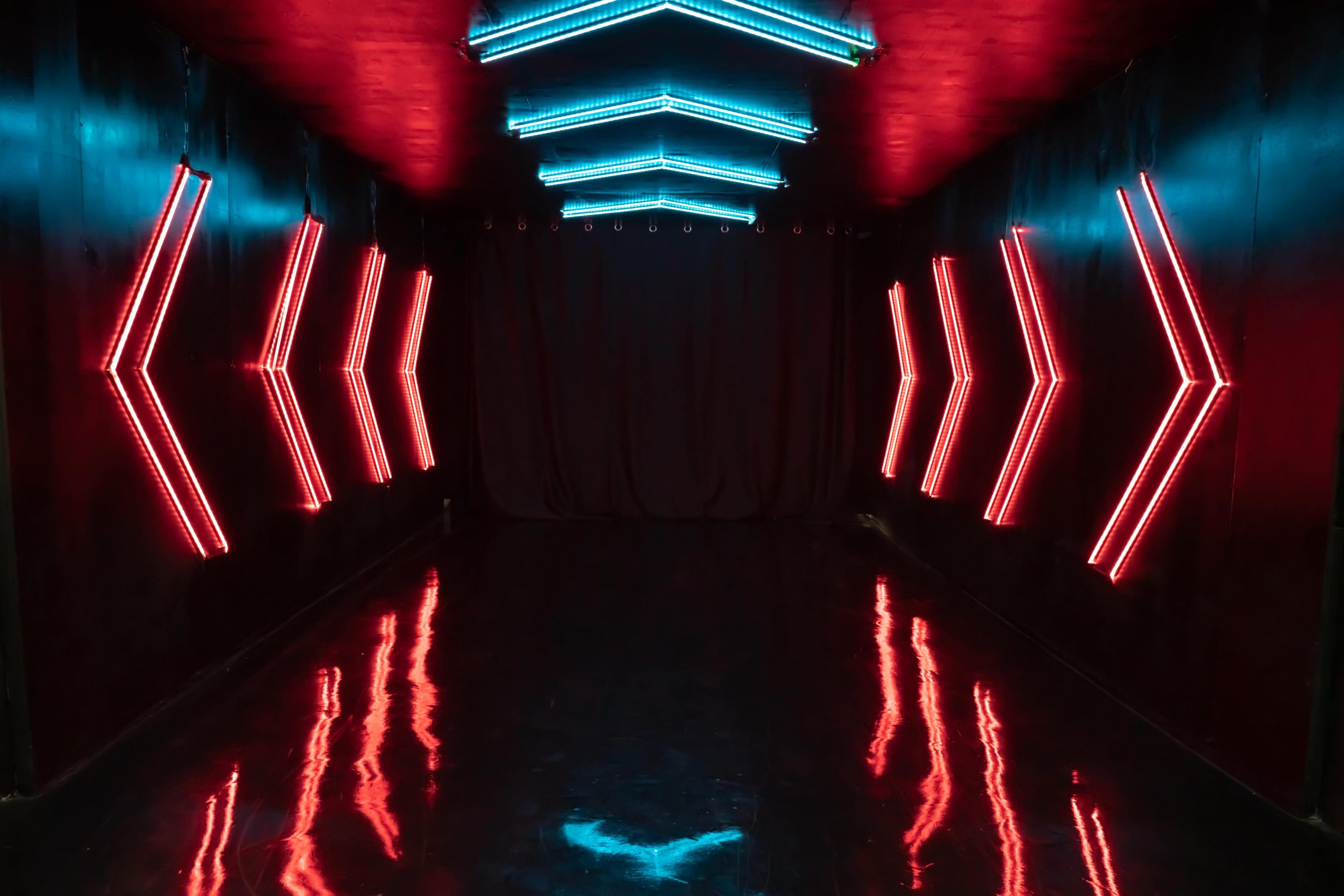 A narrow hallway lit by red and blue neon lights with a reflective floor and a black curtain at the end.
