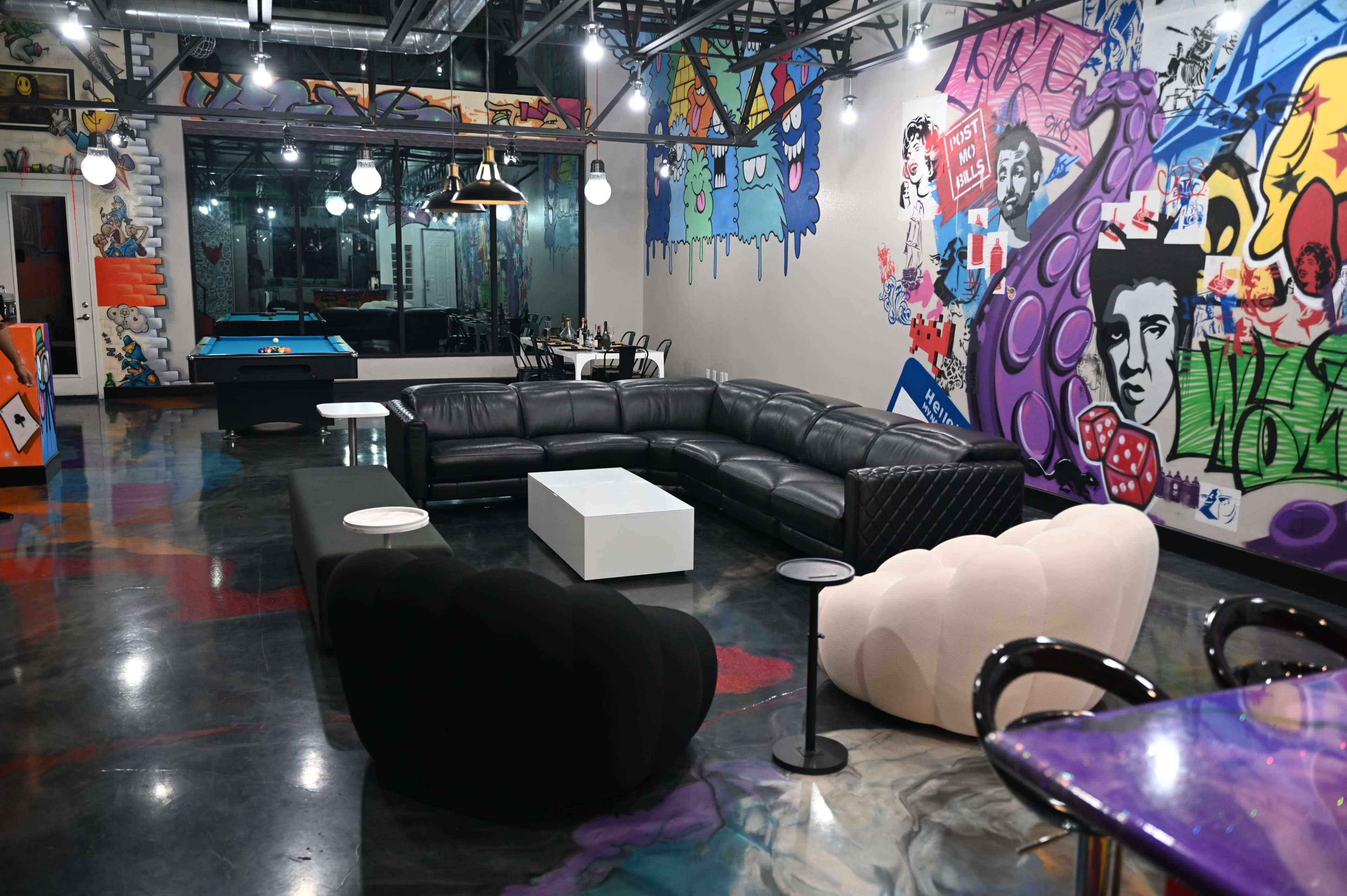 The image shows a modern lounge area featuring a sectional black leather sofa, colorful graffiti art on the walls, and various seating options, including a white table and two distinct chairs.