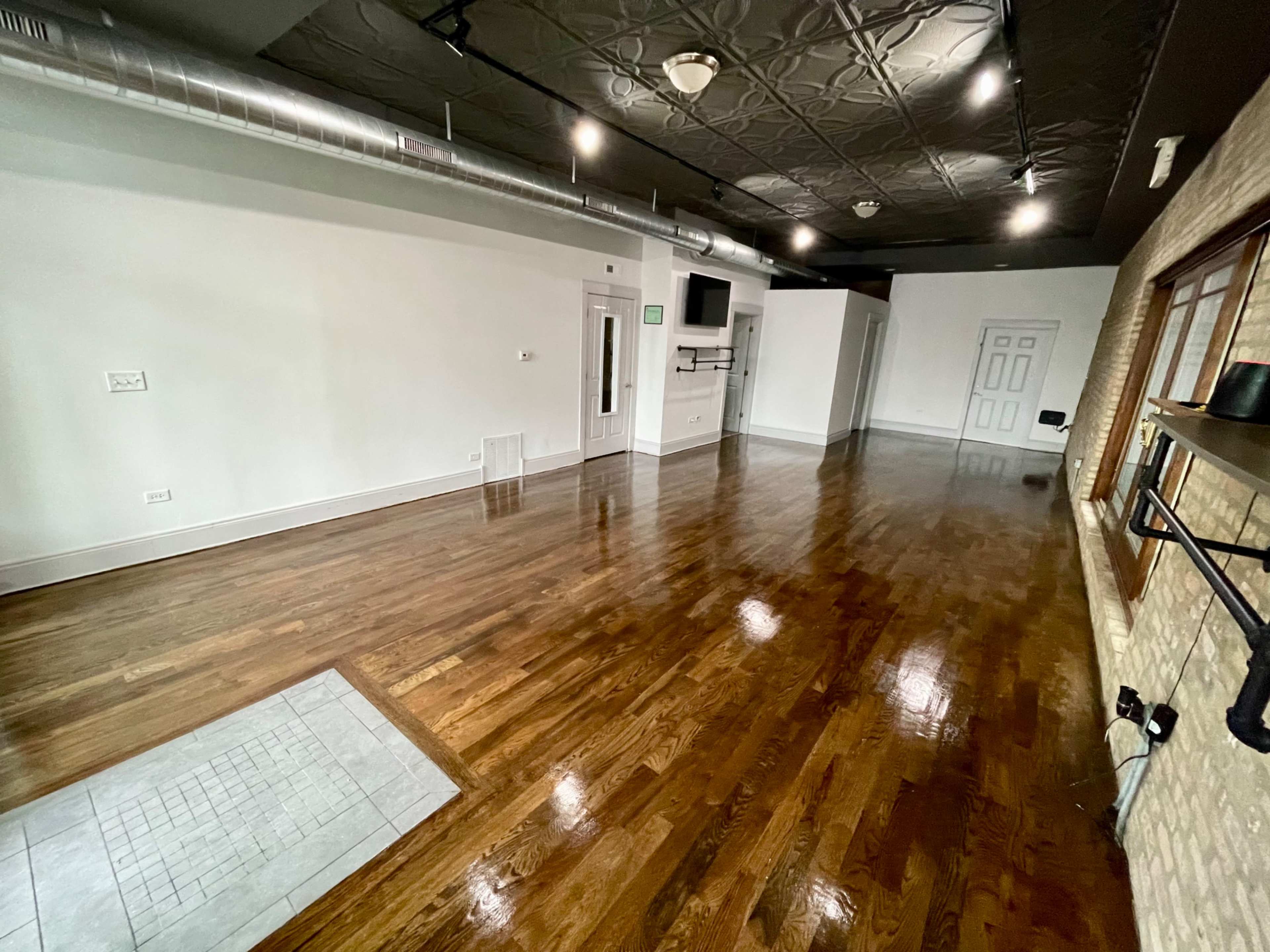A spacious room with polished hardwood floors, exposed ductwork, and a large empty area illuminated by overhead lights.