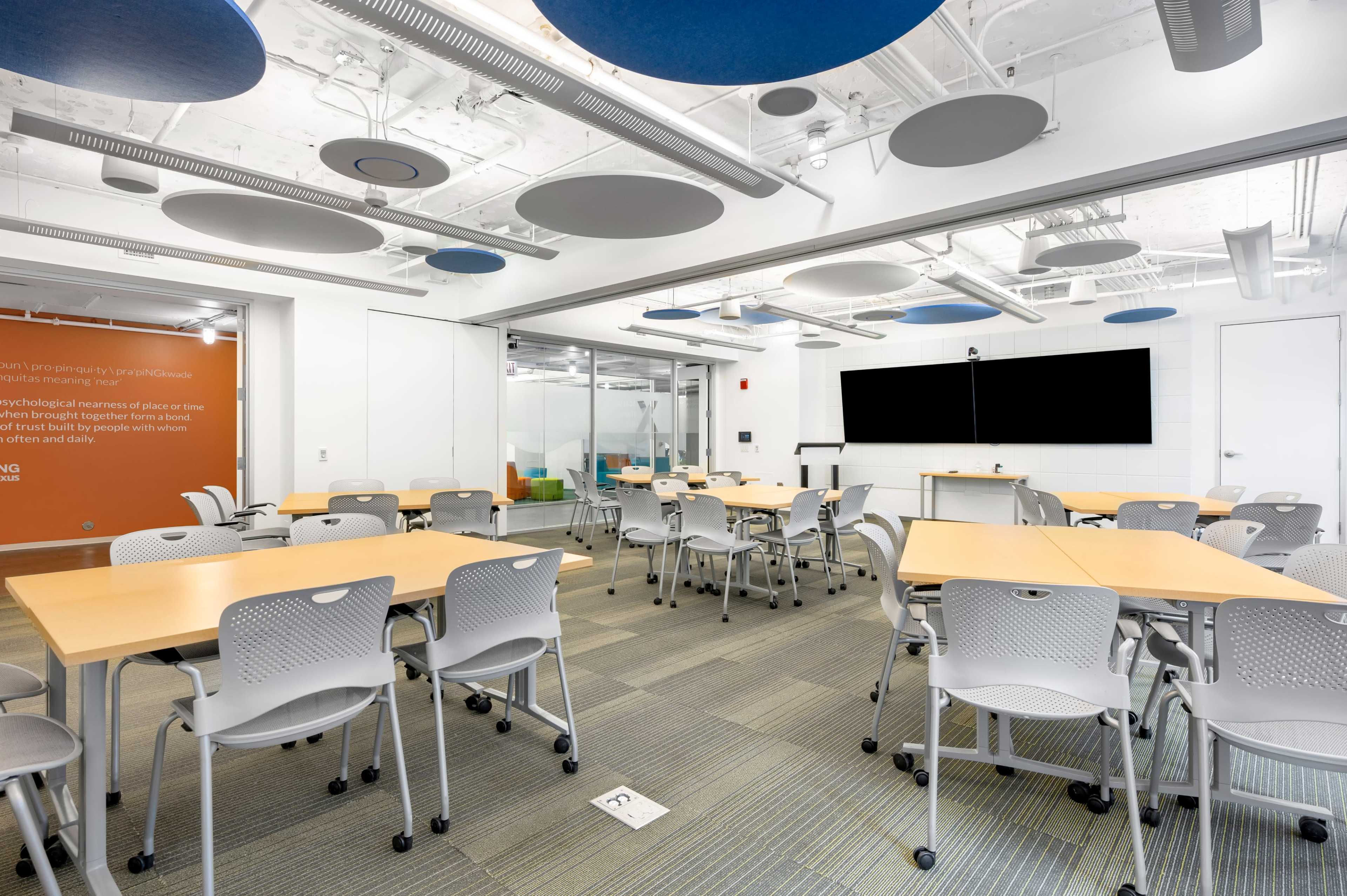 A modern classroom features several tables and chairs arranged around a large screen and glass-walled area, with acoustic panels suspended from the ceiling.