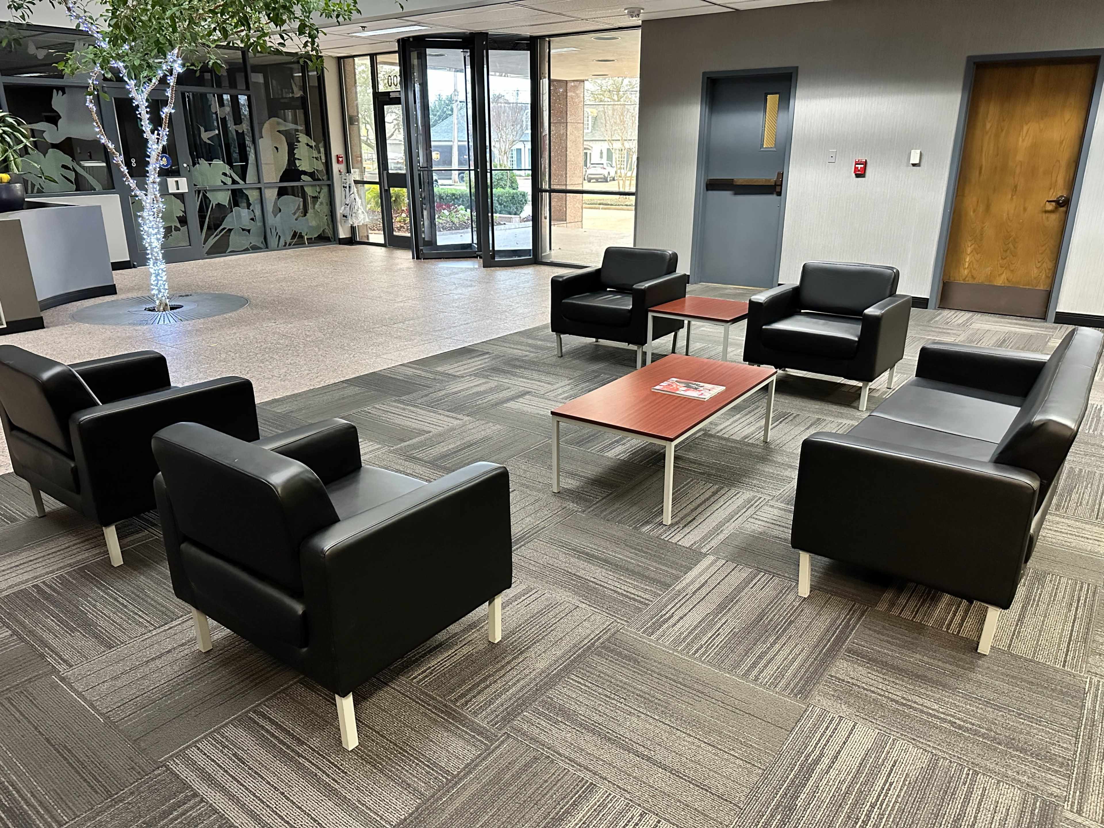 The image shows a lounge area with black leather armchairs and a small wooden coffee table arranged on a carpeted floor in front of large glass doors.